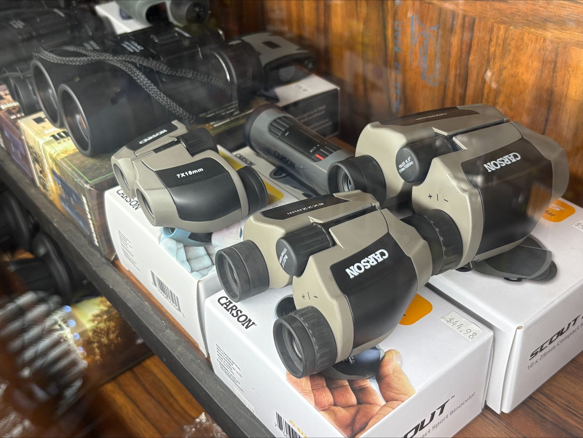 Binoculars displayed on a shelf, various brands and sizes, mostly in black and tan. Some in boxes, store setting.