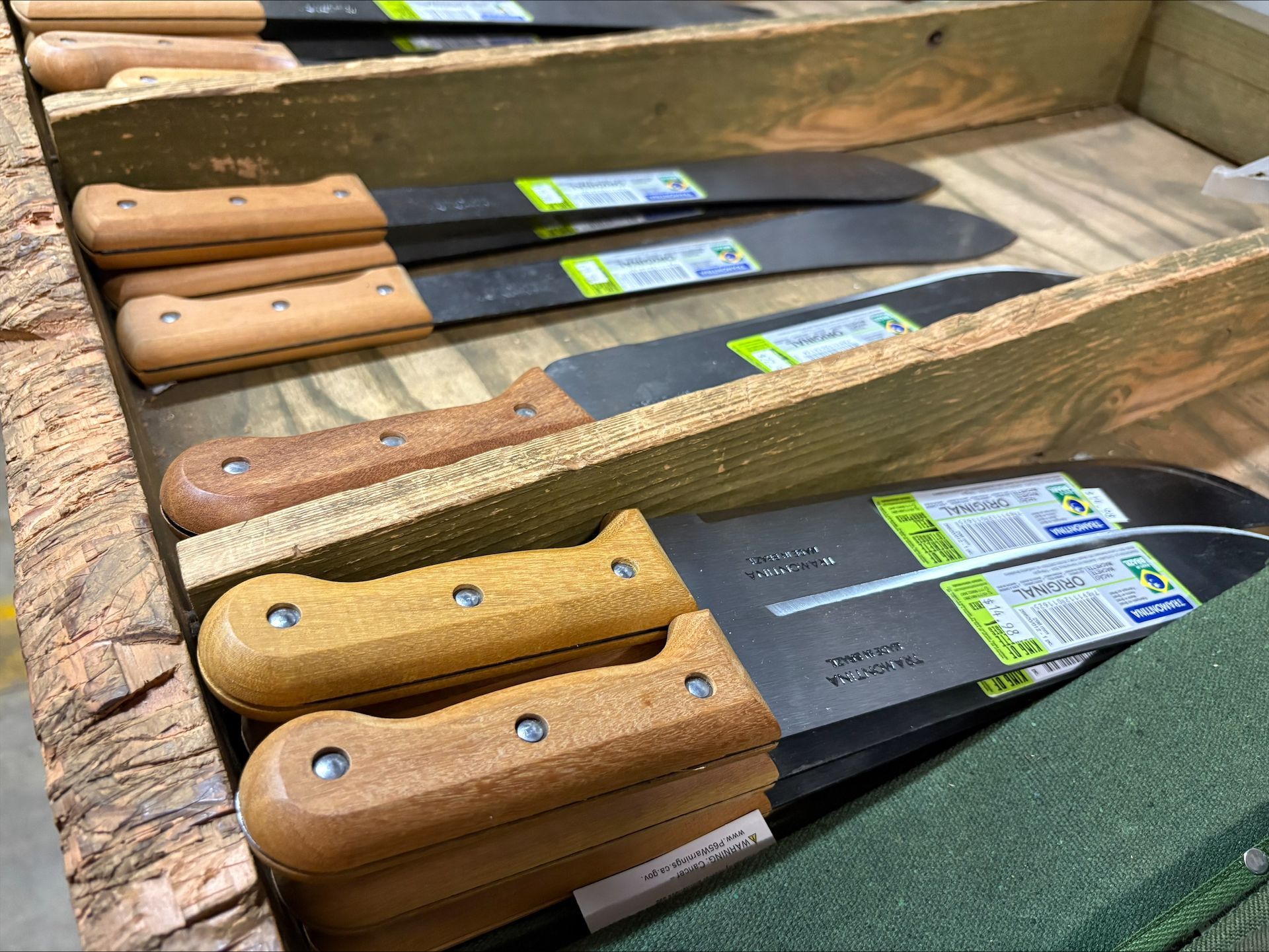 Machetes with wooden handles displayed in a wooden bin. Black blades with product labels.