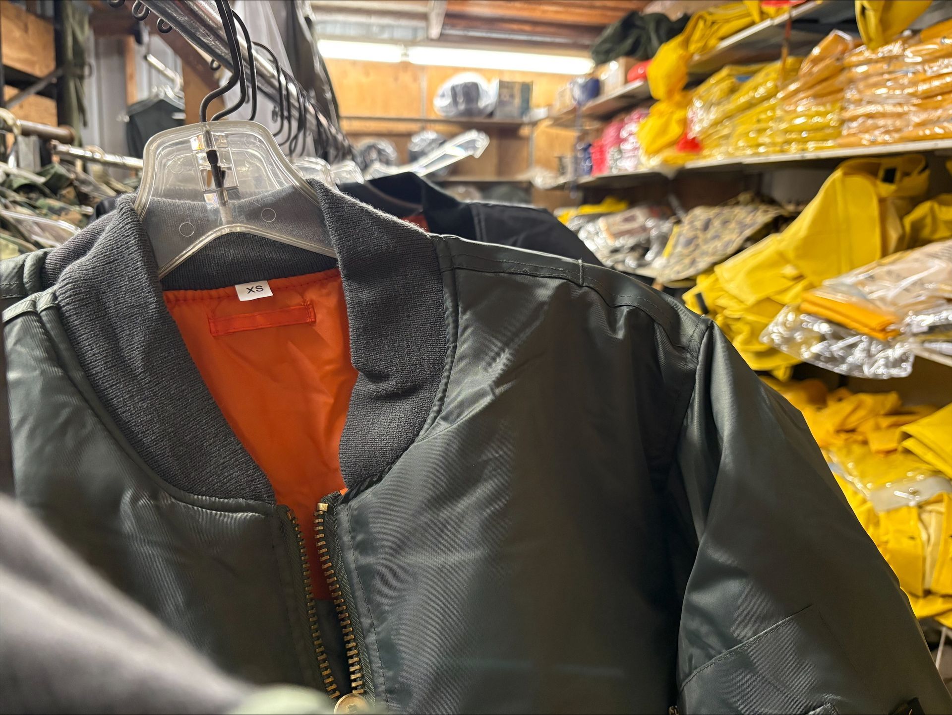 Green bomber jacket with orange lining hanging in a store. Shelves with yellow items are in the background.