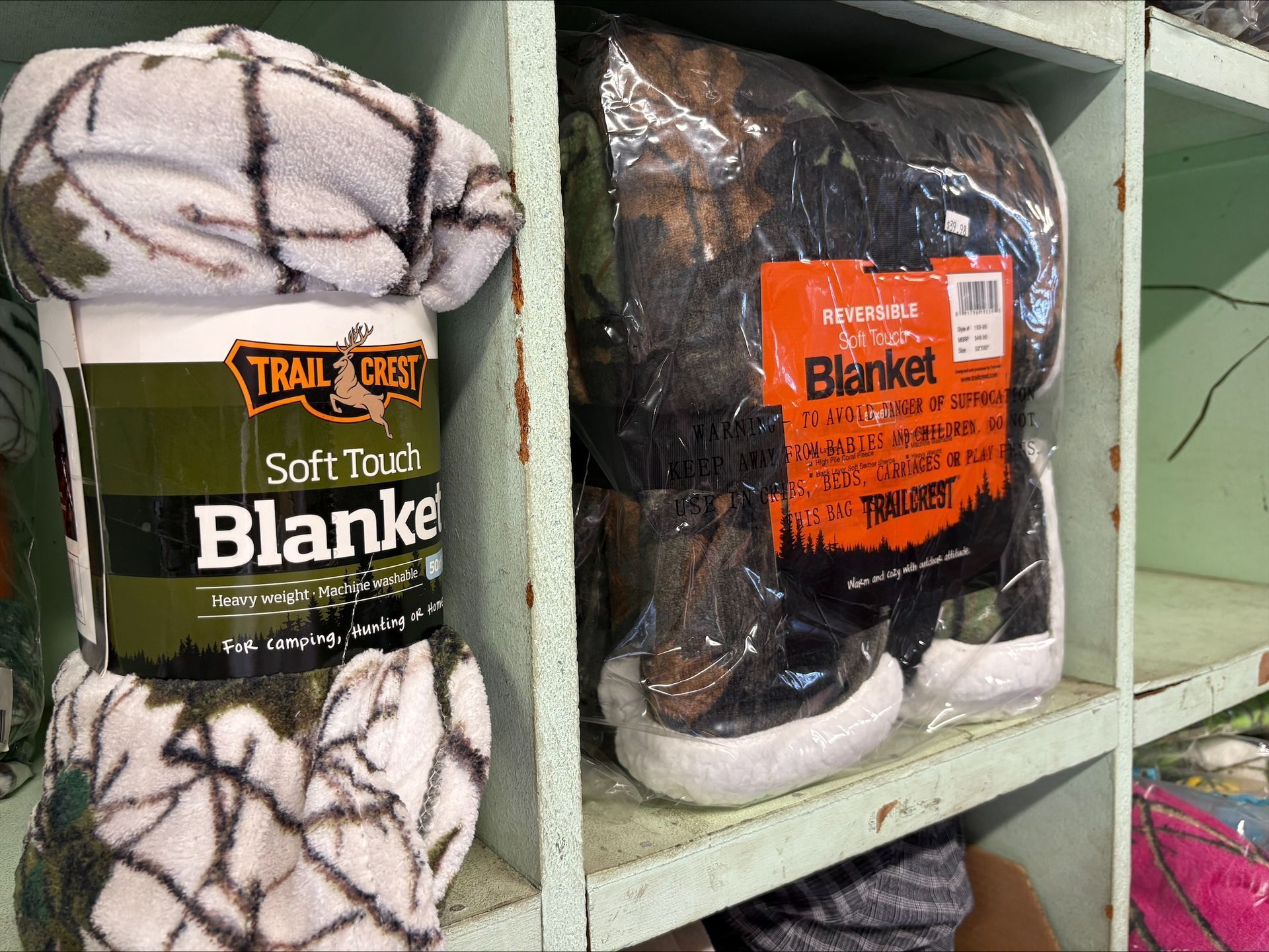 Blankets in camo patterns displayed on a shelf. One is rolled, and one is packaged.