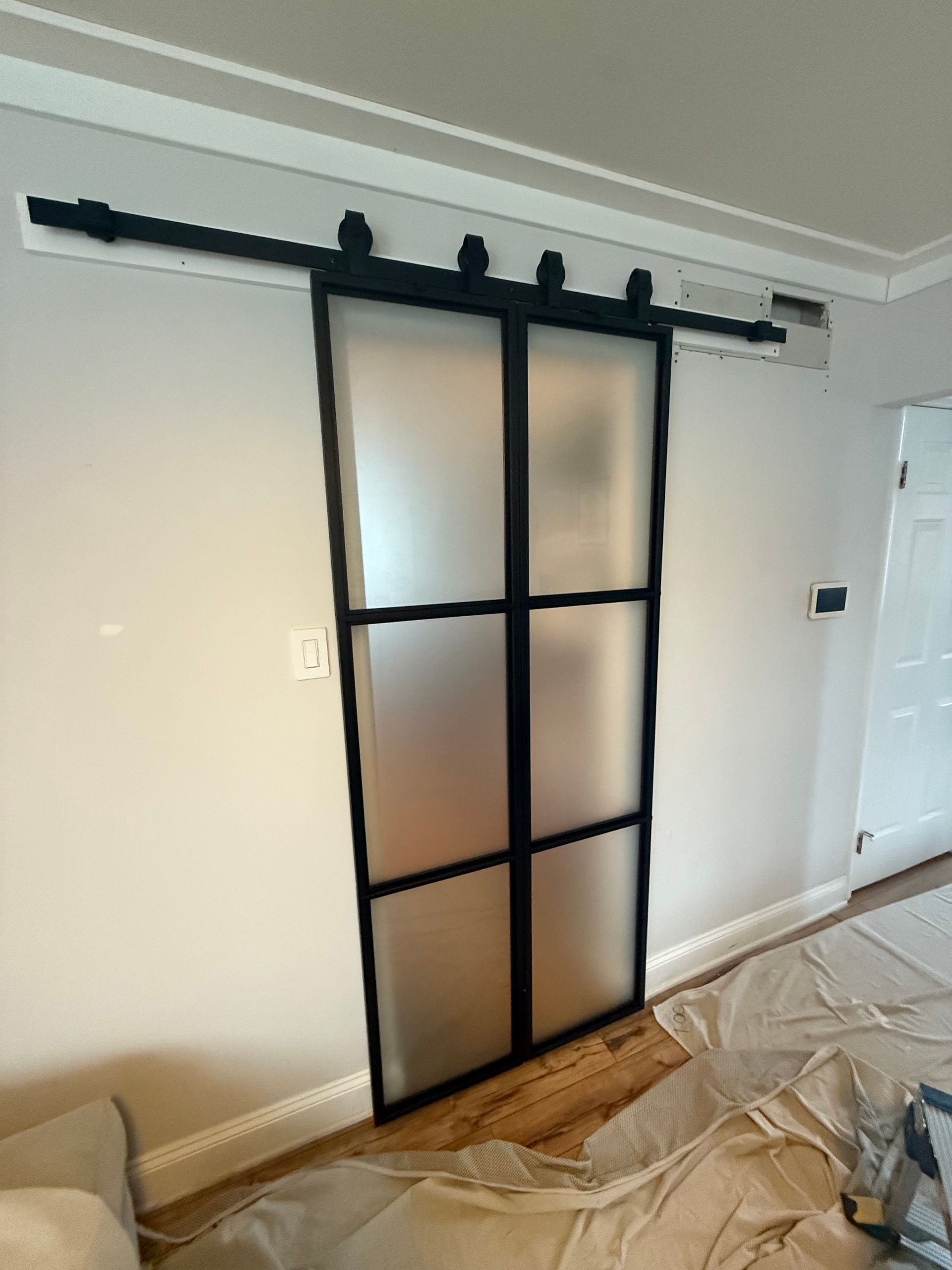 Sliding black-framed frosted glass door on a track, mounted on a white wall above wooden flooring.