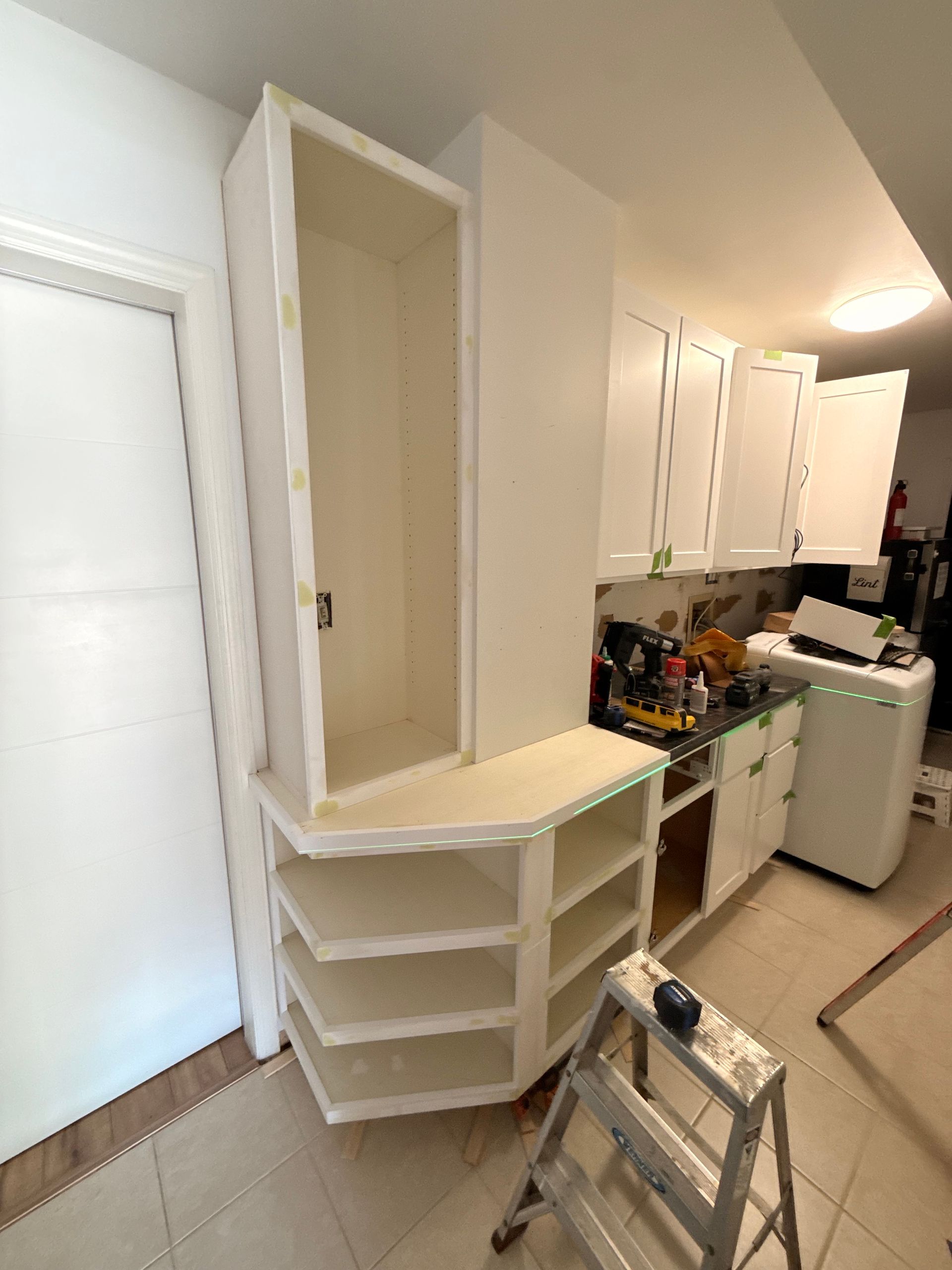 Kitchen cabinets being installed, unfinished white cabinets and shelving units. Ladder in front.