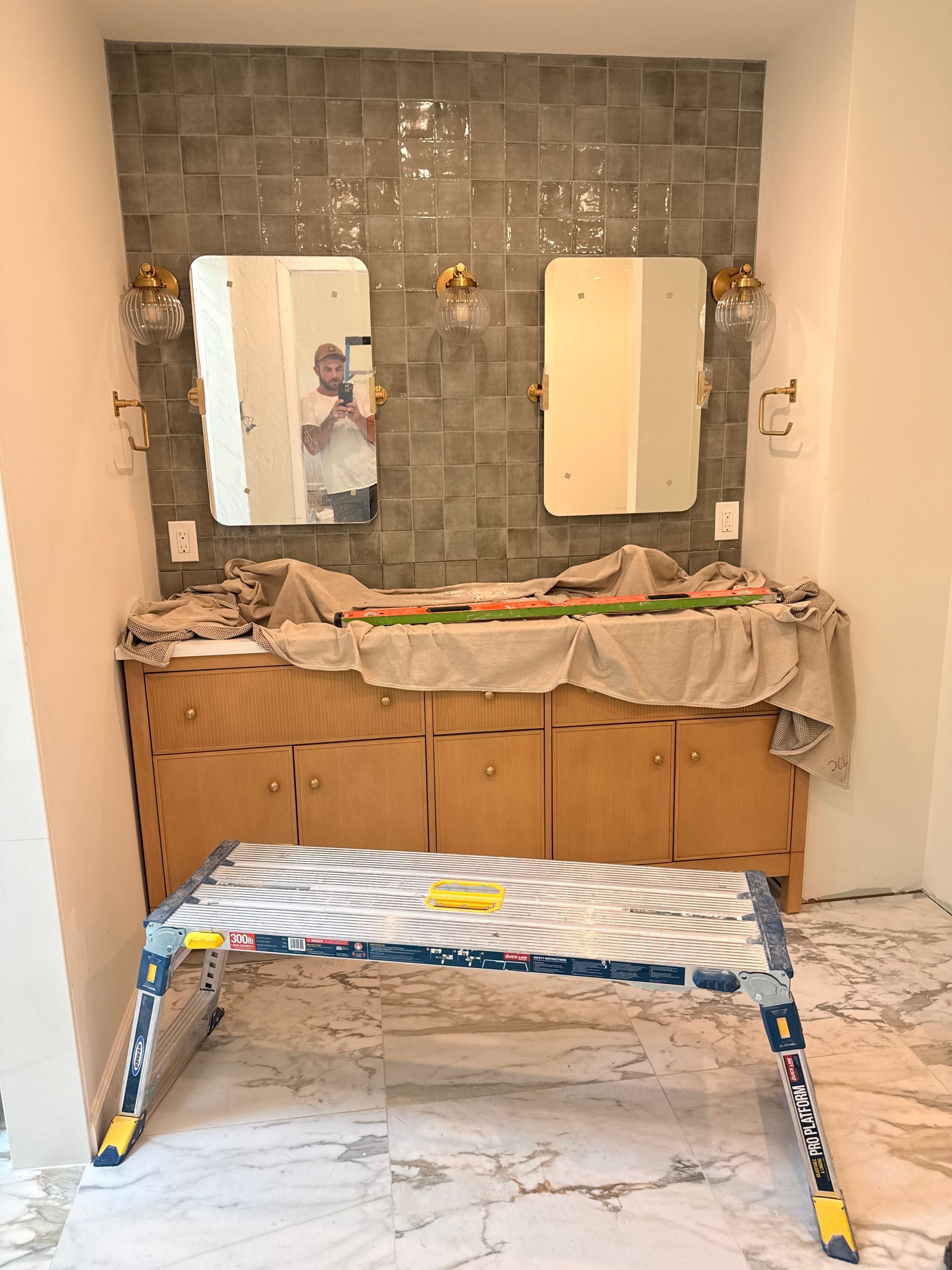 Bathroom under construction: wood vanity, tile backsplash, two mirrors, step stool. Person reflected in mirror.
