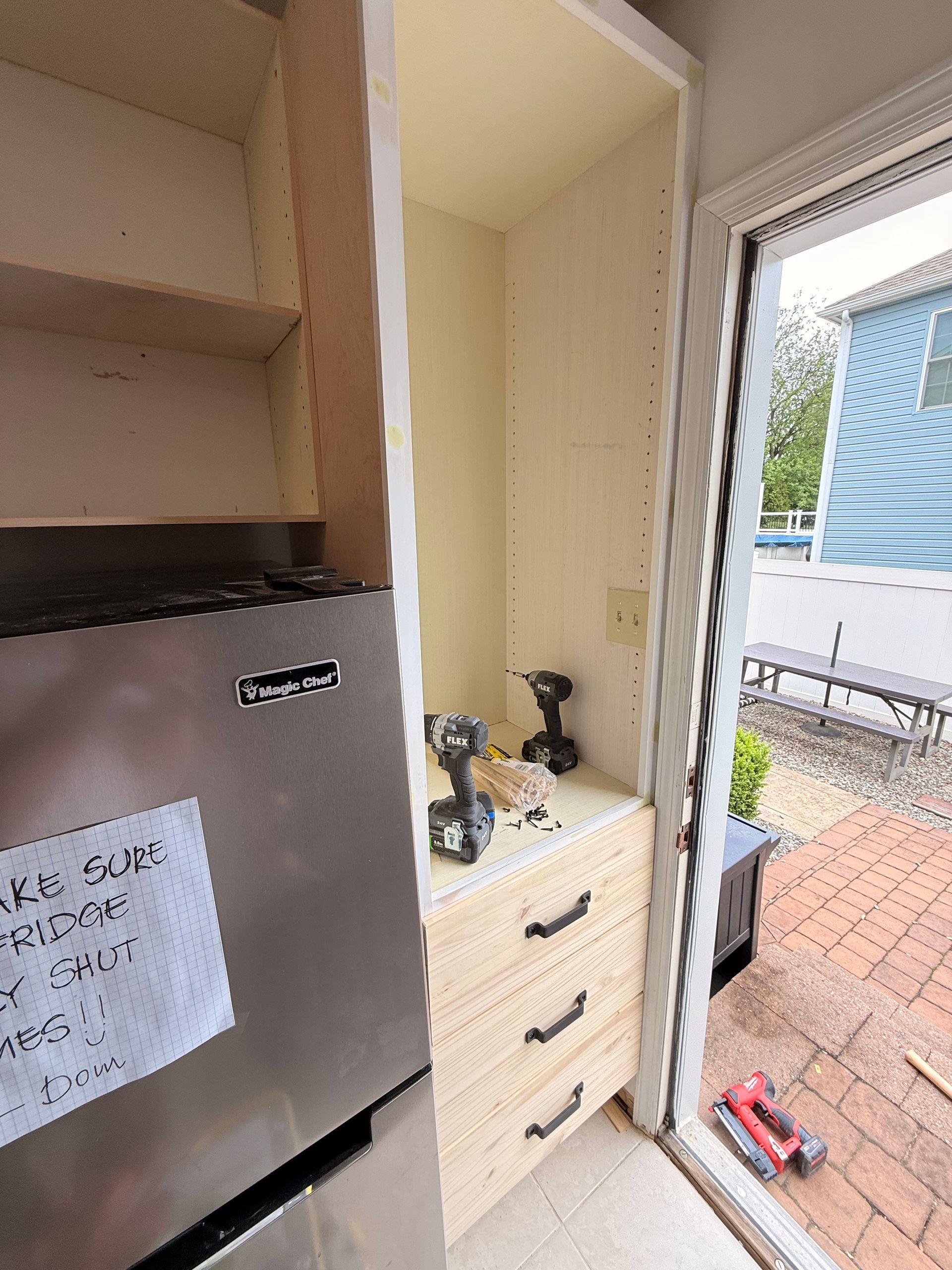 Kitchen cabinet under construction near a sliding glass door and refrigerator. Beige cabinetry, black hardware, power tools visible.