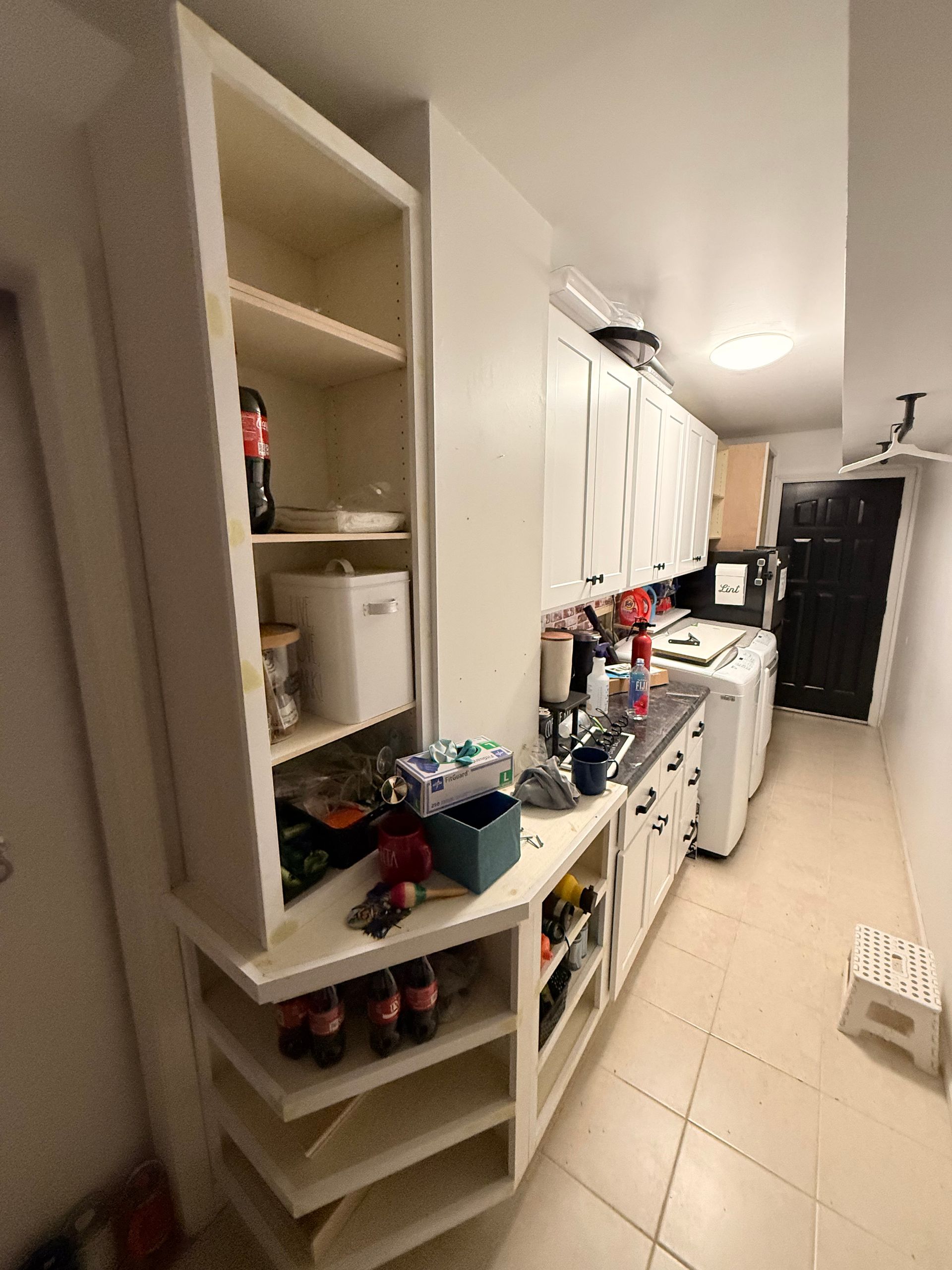 A narrow kitchen with white cabinets, shelves, and appliances. Shelving unit on the left.