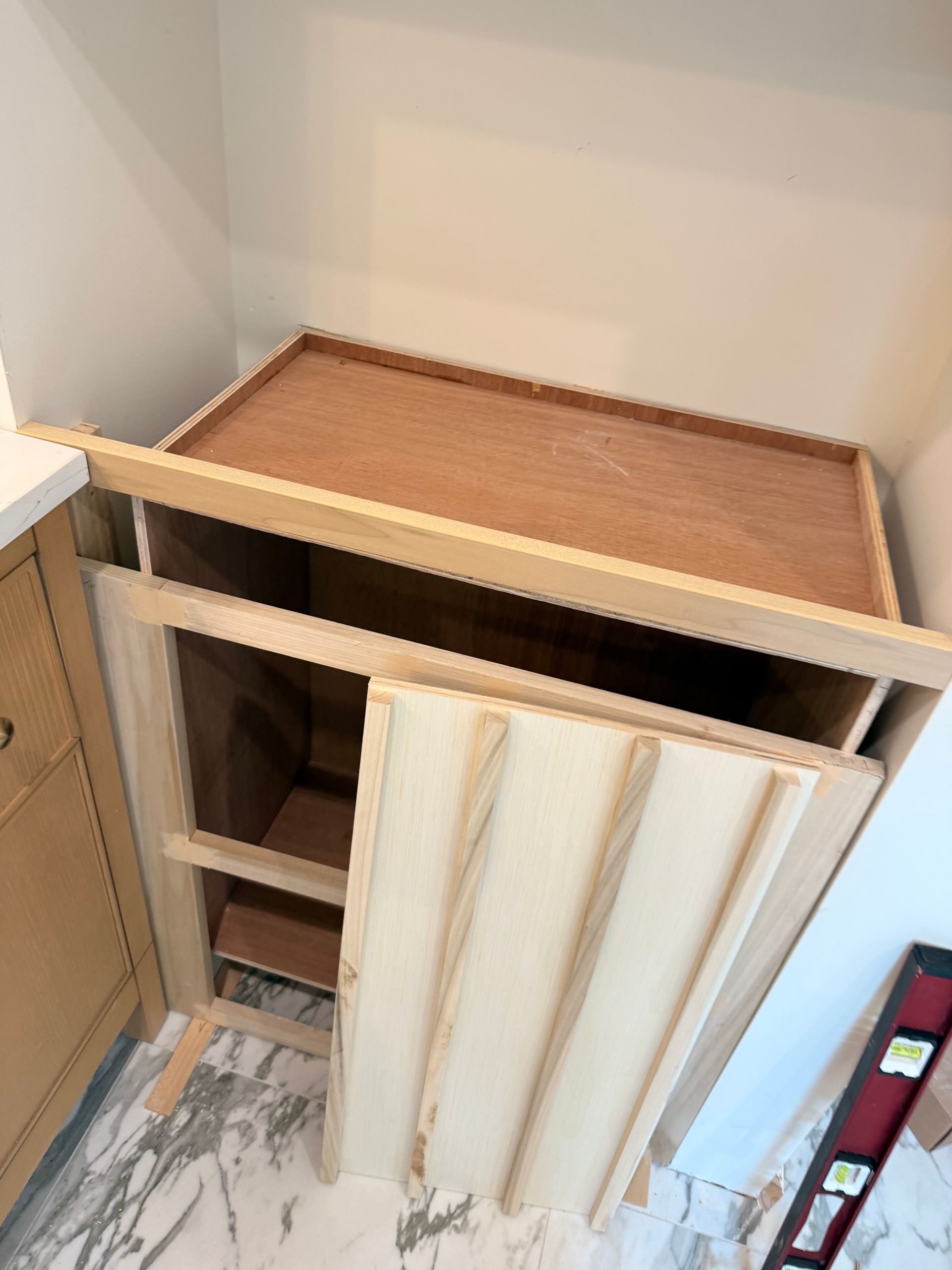 Wooden cabinet under construction; interior shelving visible, door off. Light-colored wood, white walls, and marble floor.