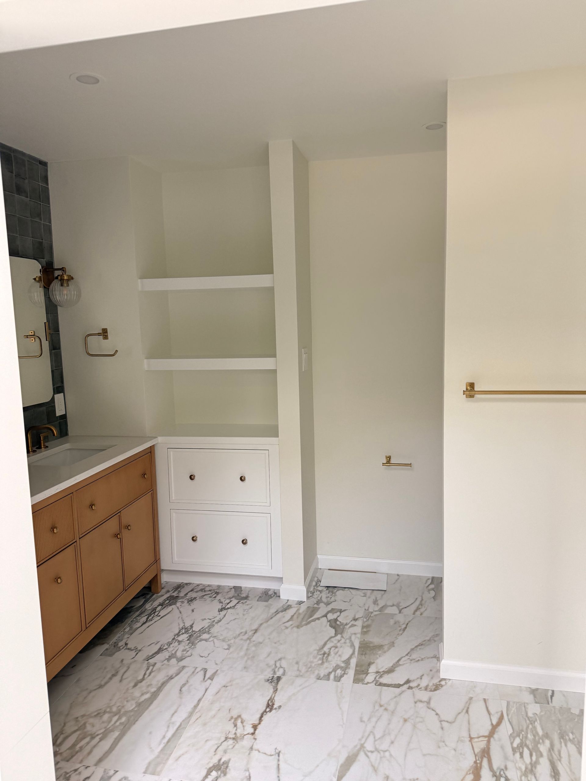 Bathroom with a marble floor, wood vanity, white shelving, and brass towel rack.