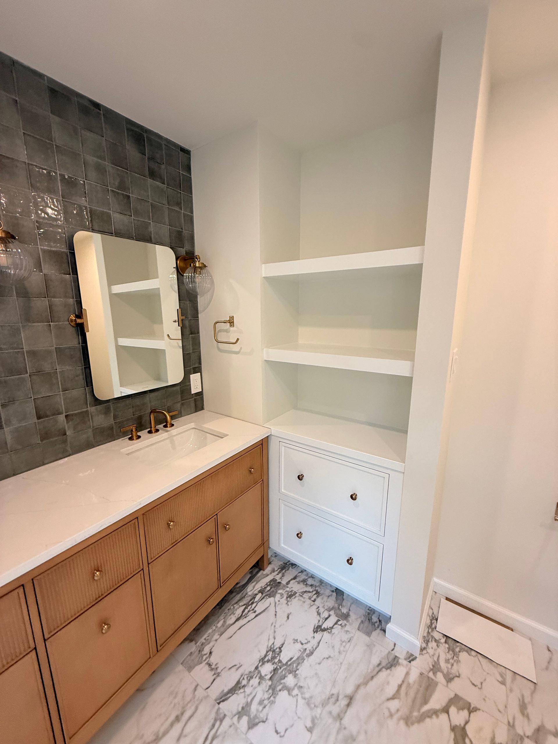 Bathroom with wood vanity, marble floor, white shelving, and dark tile accent wall.