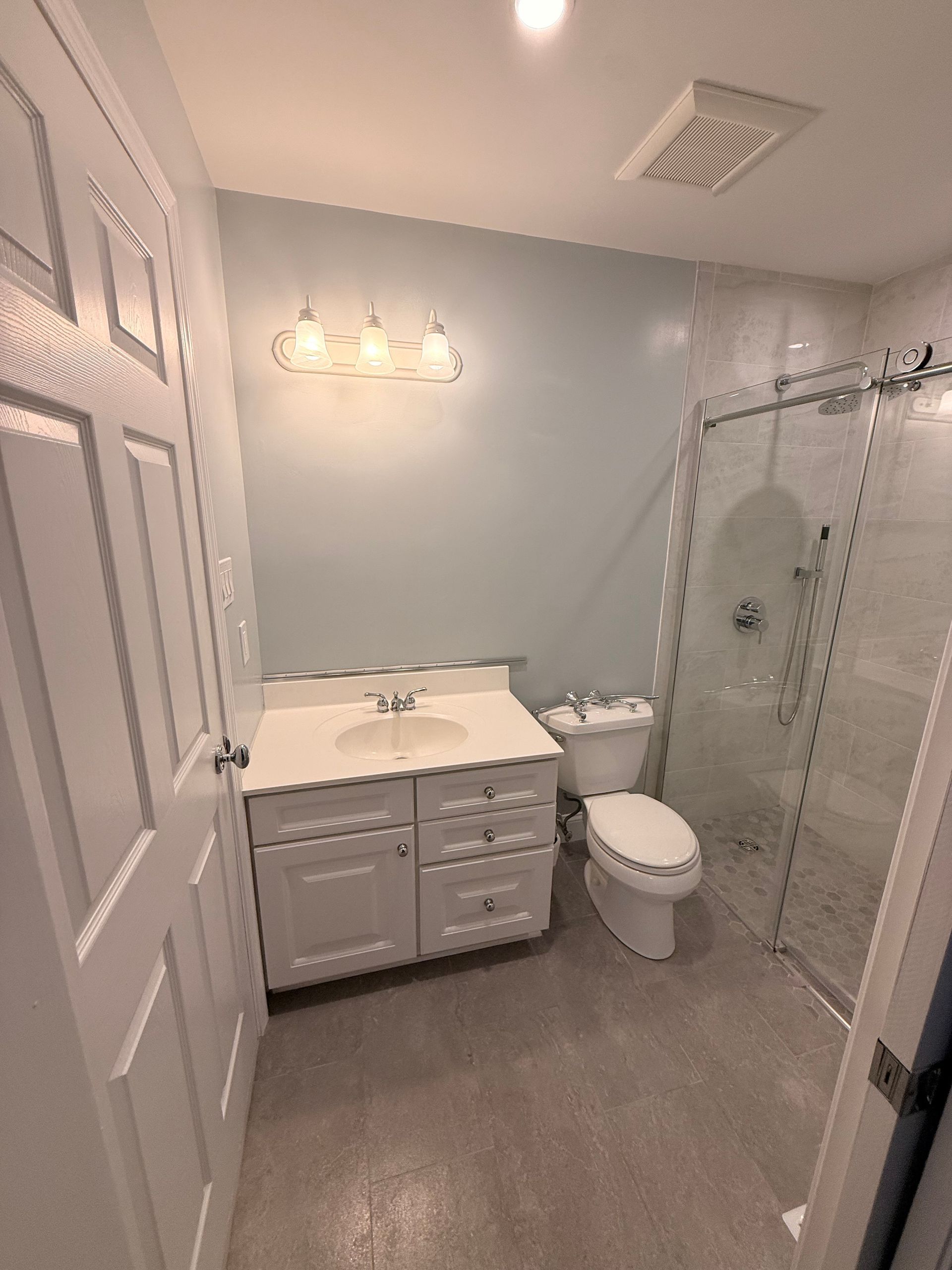 Bathroom with white vanity, toilet, and shower. Light blue wall, gray floor.