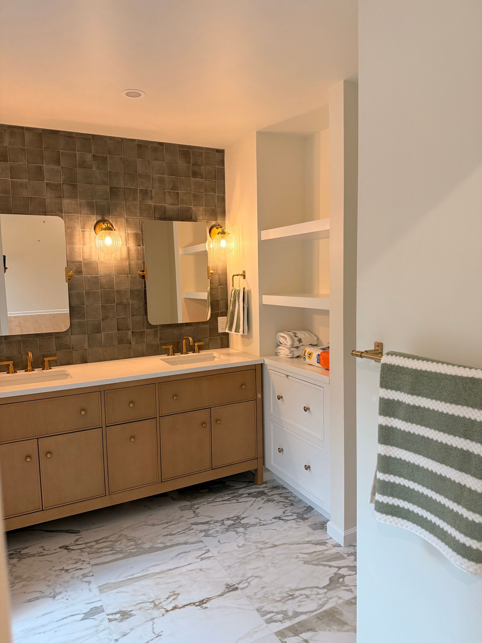 Bathroom with light wood vanity, square tile wall, and built-in shelving. A towel hangs on the door.