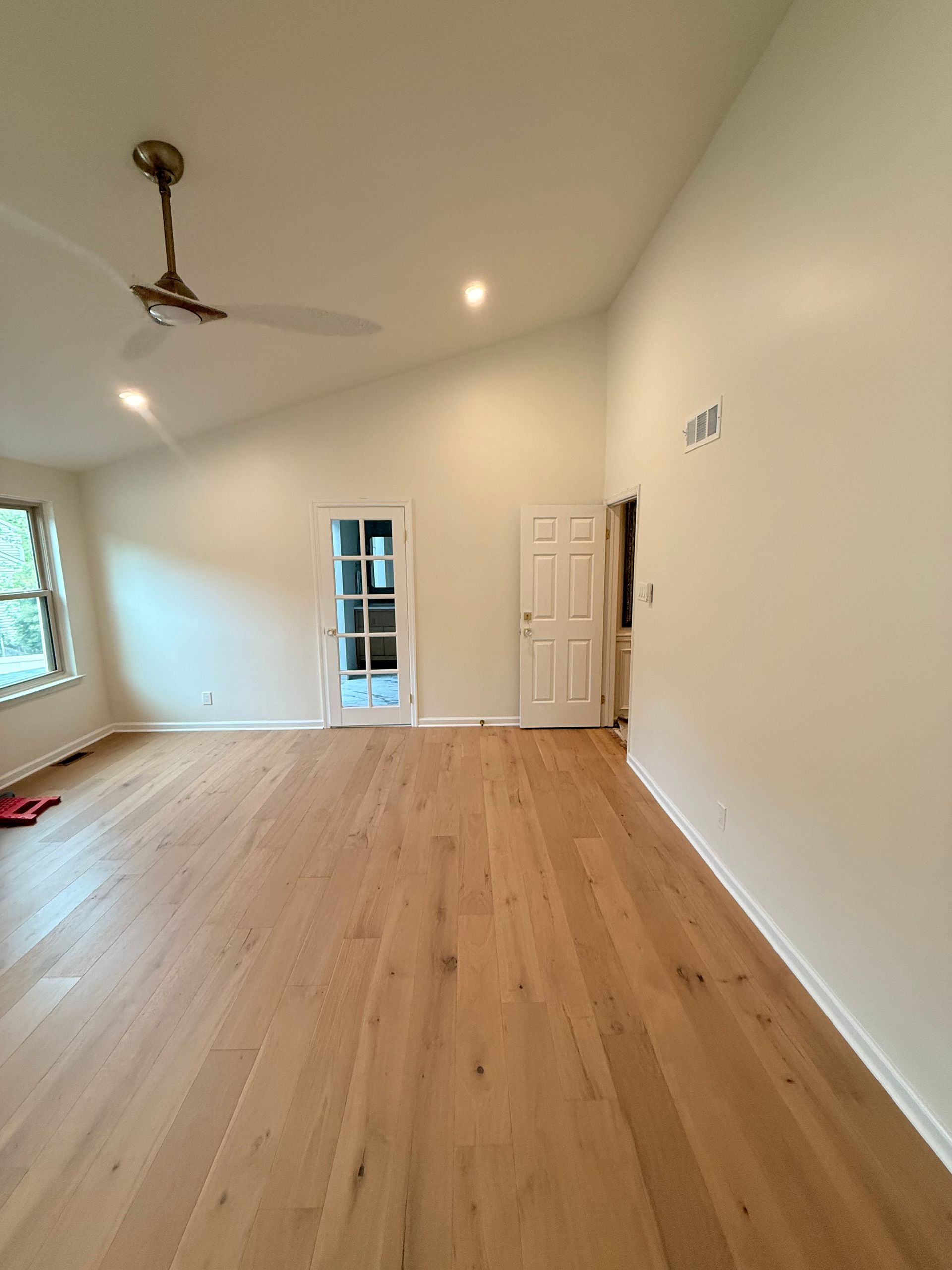 A bright, empty living room with wood flooring, white walls, and a ceiling fan. Two doorways and a window are visible.
