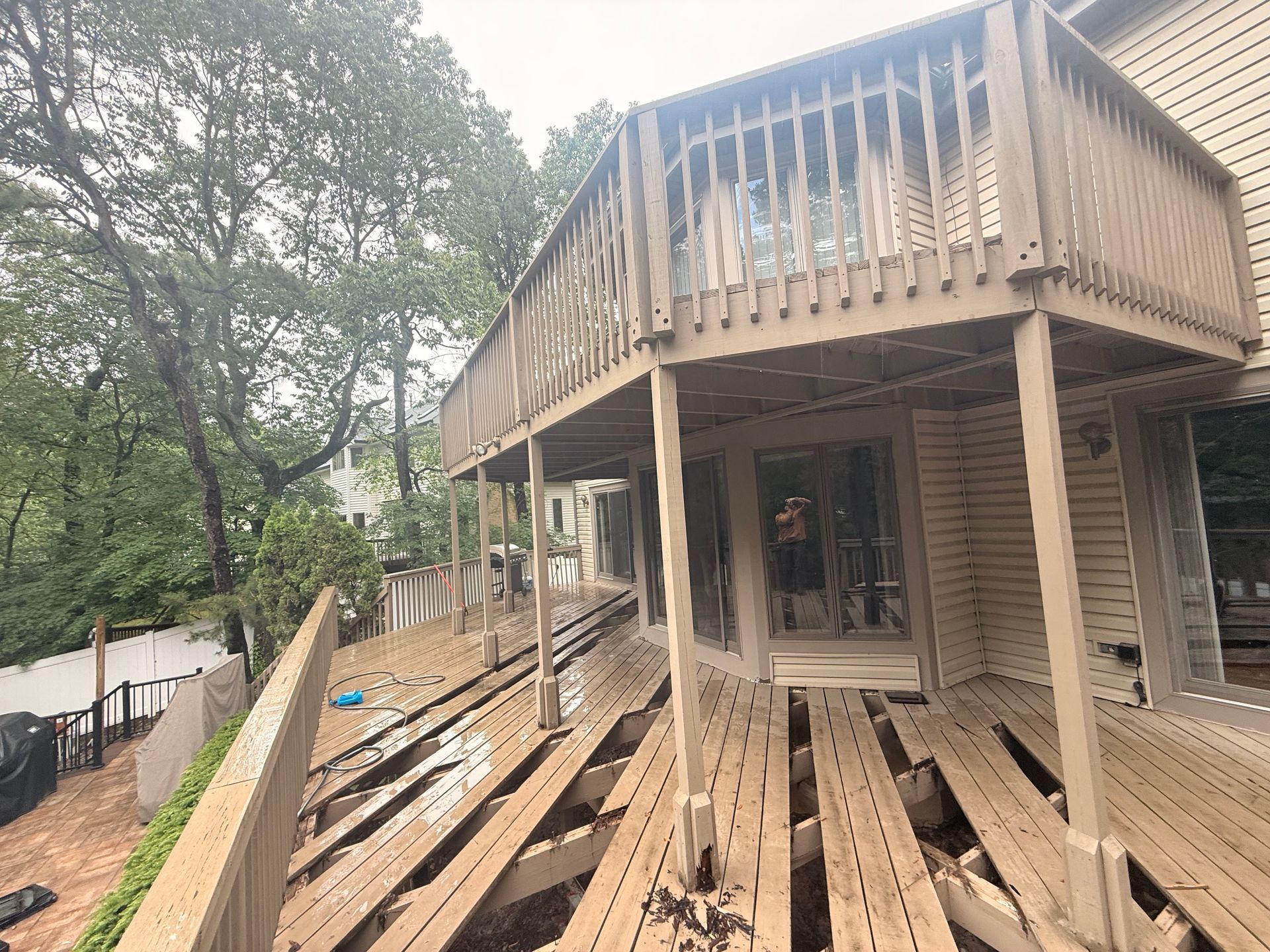 Damaged two-story wooden deck with missing boards, overlooking a yard with trees.