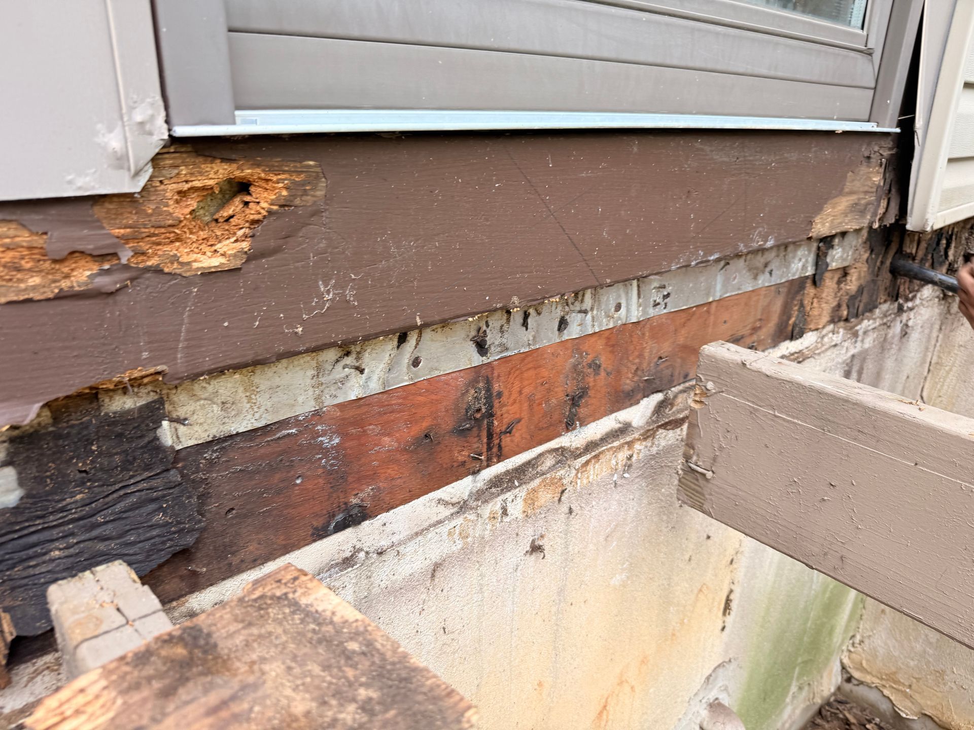 Exterior house wall with damaged siding and exposed wood, revealing brick and concrete foundation. Brown and beige tones.