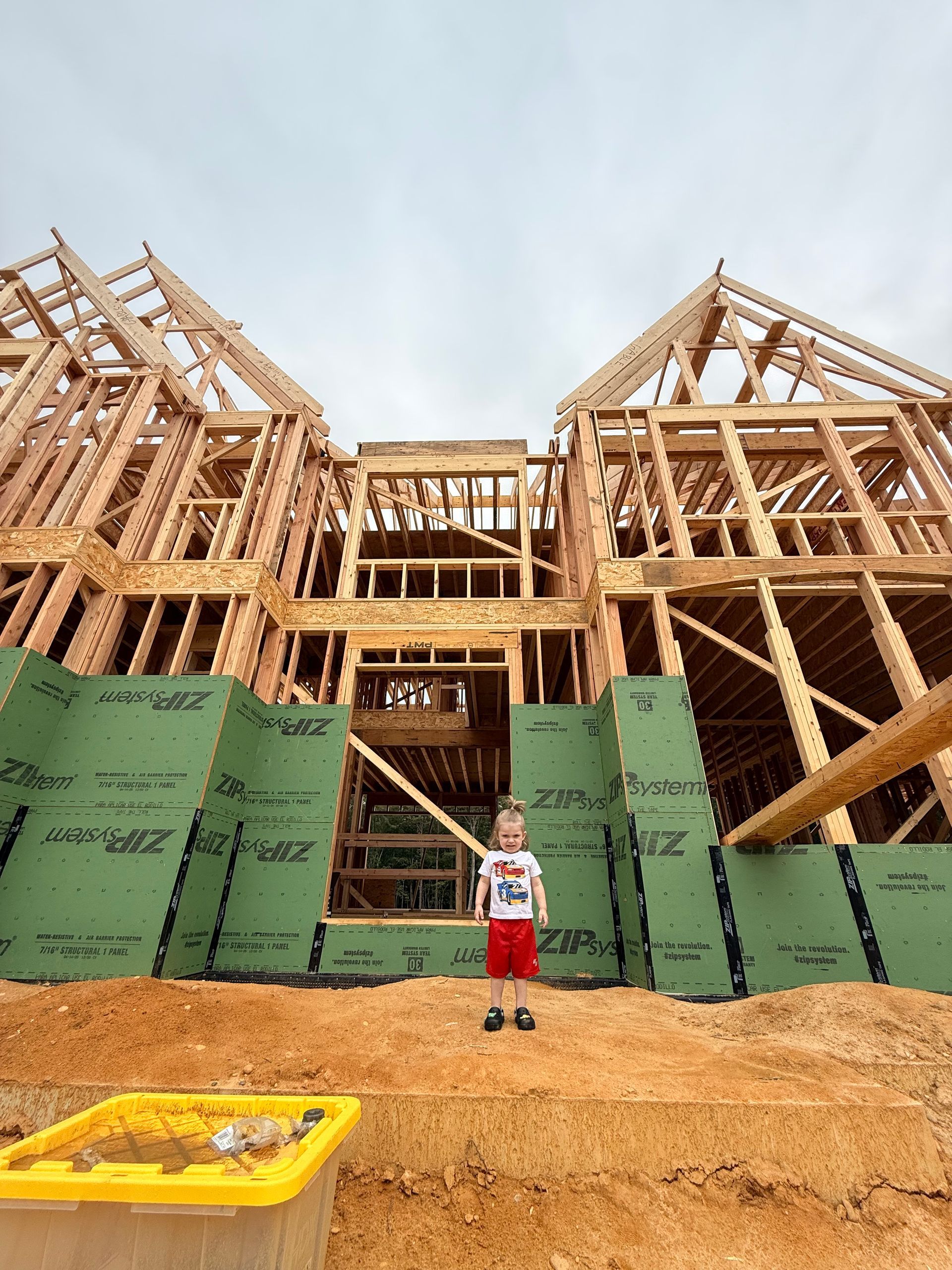 Child stands in front of a house under construction. Wooden frame, green panels, and overcast sky.
