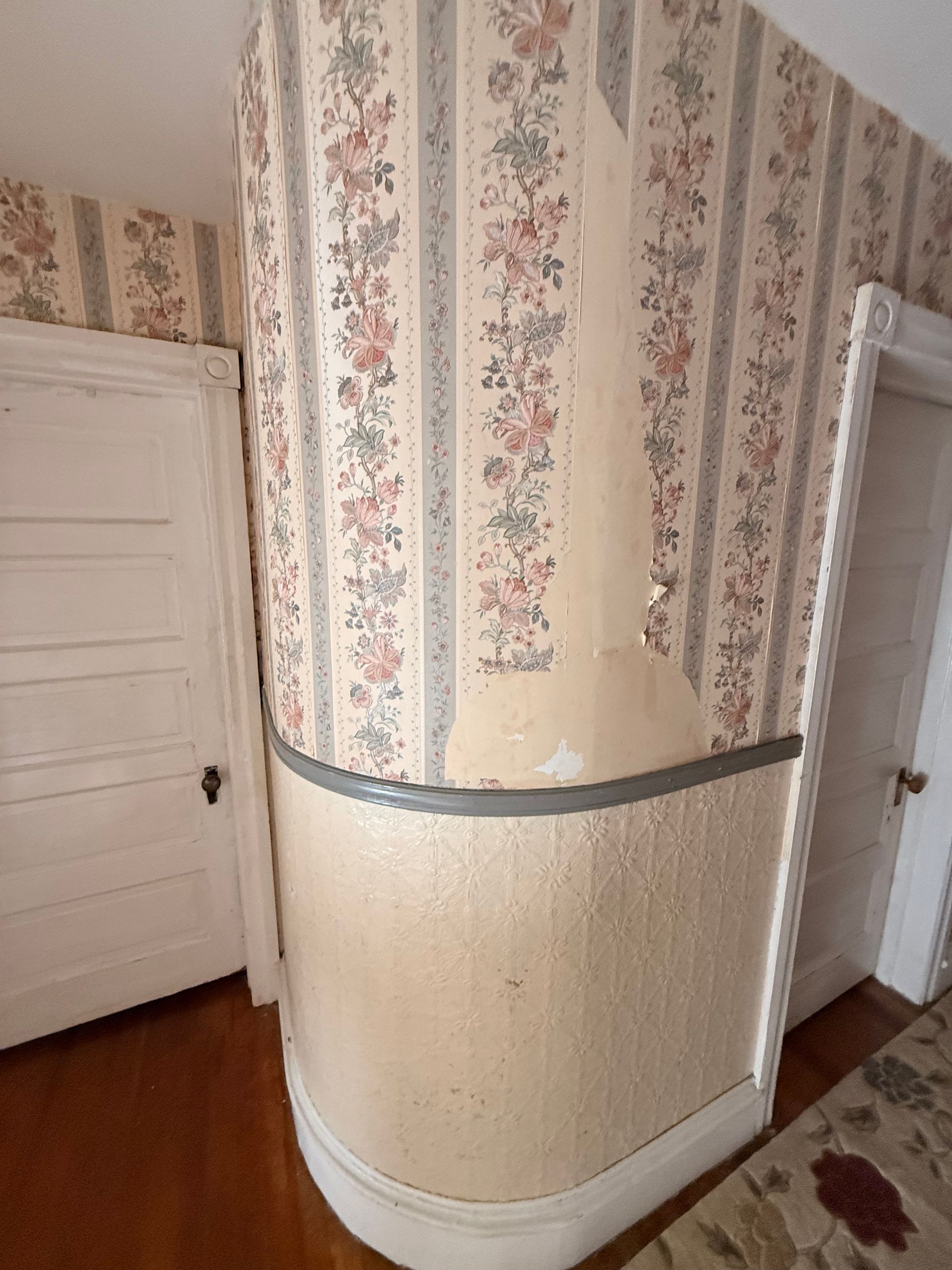 Room with curved wall, floral wallpaper peeling, showing the plaster beneath. Two white doors.