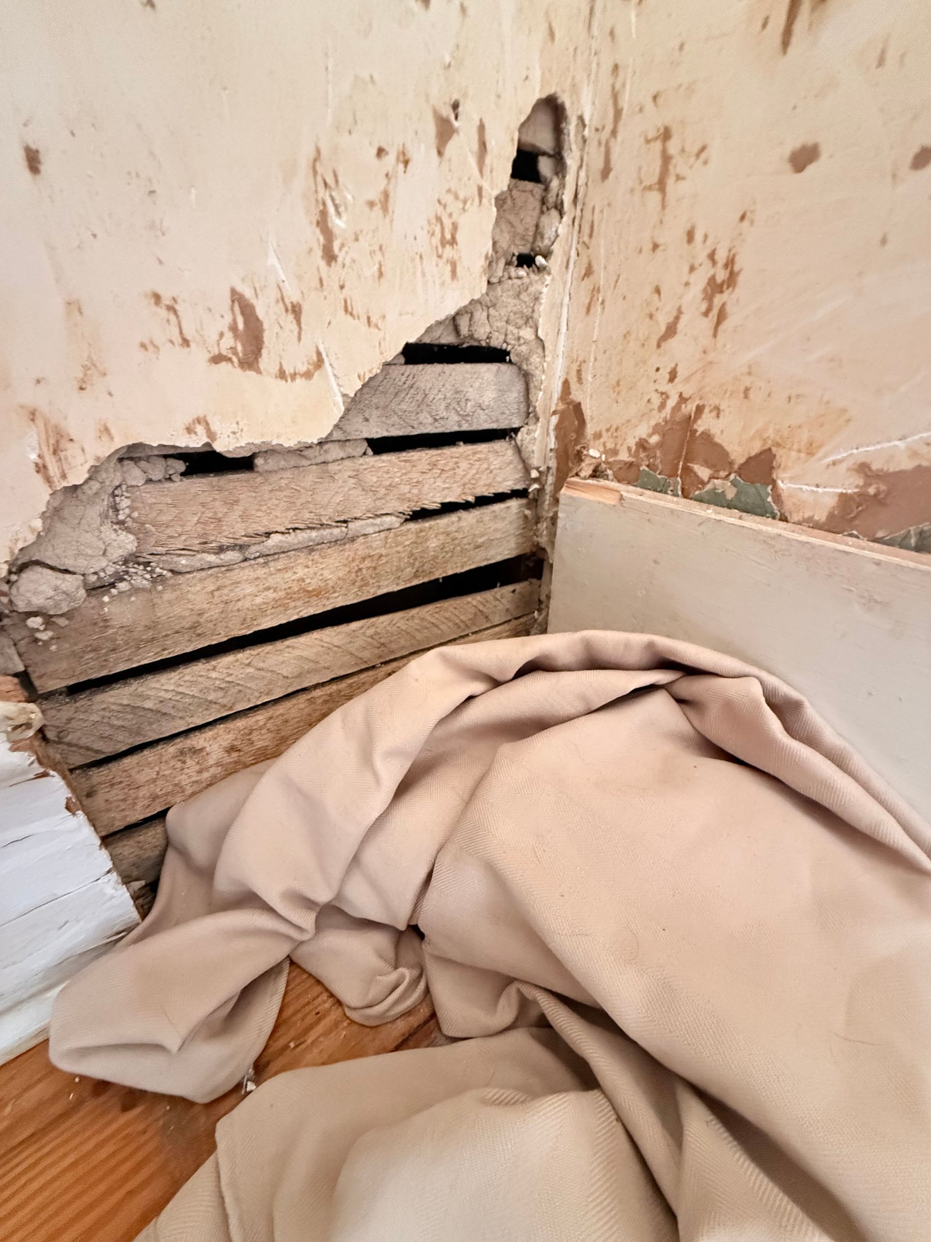 Hole in wall reveals wooden slats. Beige cloth on floor. White trim and damaged plaster.