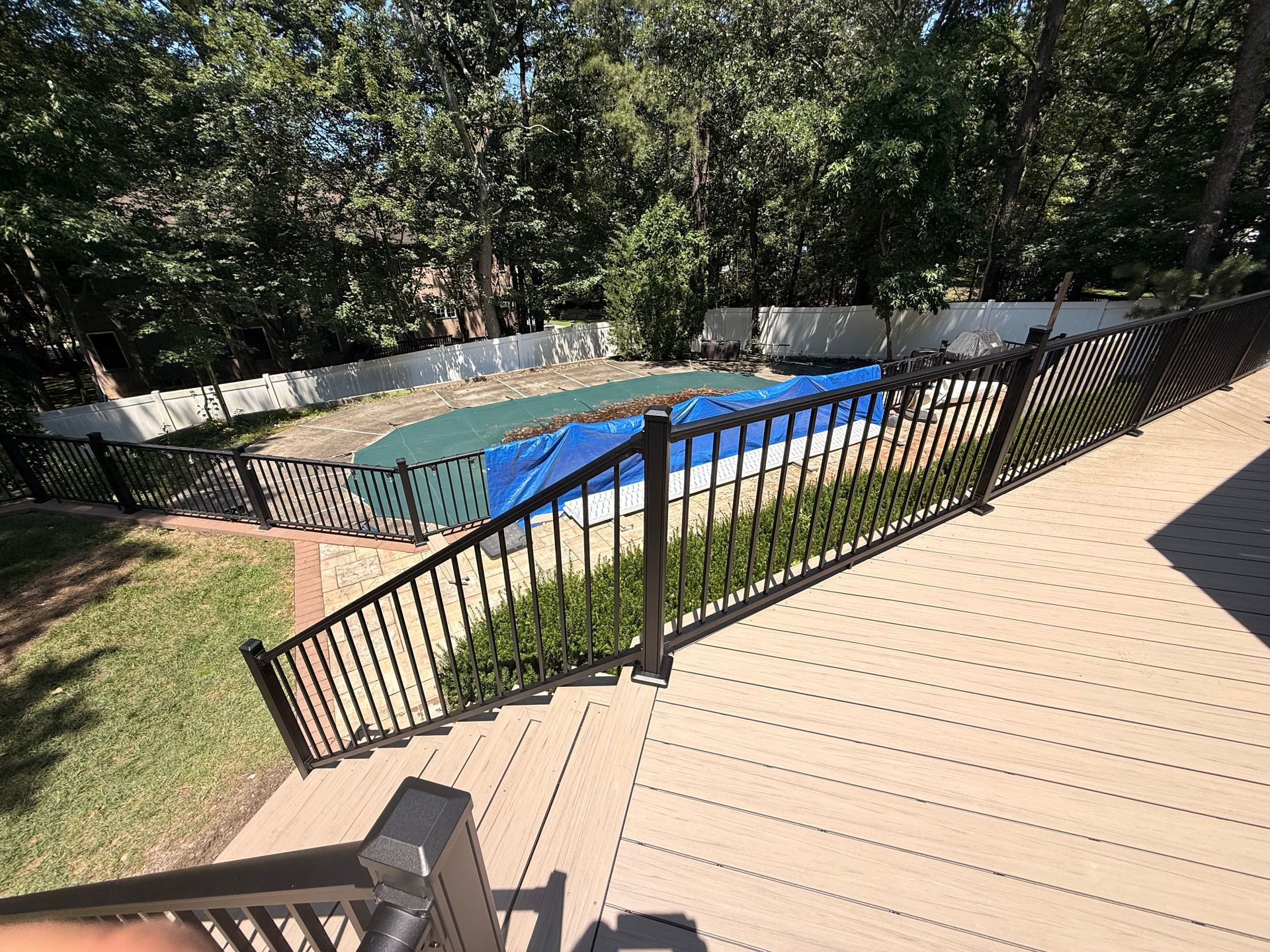 A brown deck with a black fenced-in pool covered by a blue and green tarp. Trees in the background.