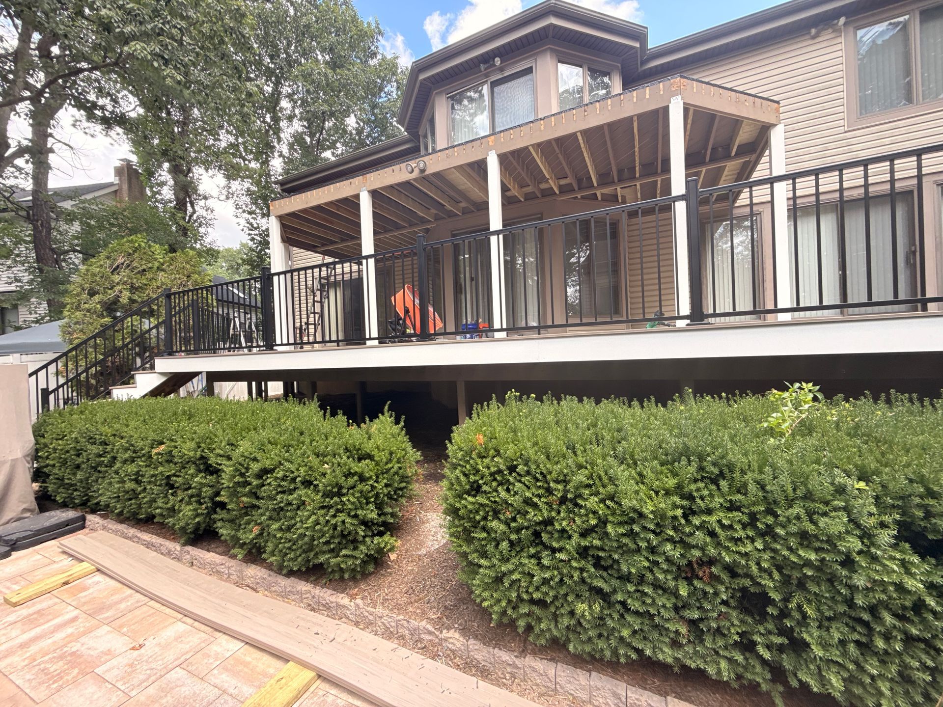 Back of a two-story house with a wooden deck, bushes in front. Brown, white, black, and green colors.