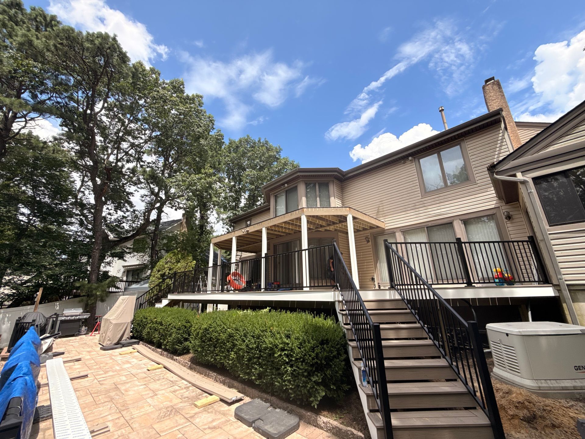 Two-story house with wooden deck, stairs, and bushes. Blue sky with clouds. Brick patio in the foreground.