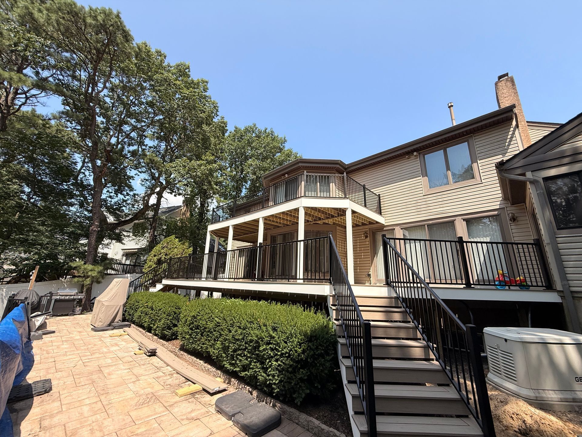 Two-story house with large deck, steps, and black railing. Blue sky, green trees, and shrubs are also visible.