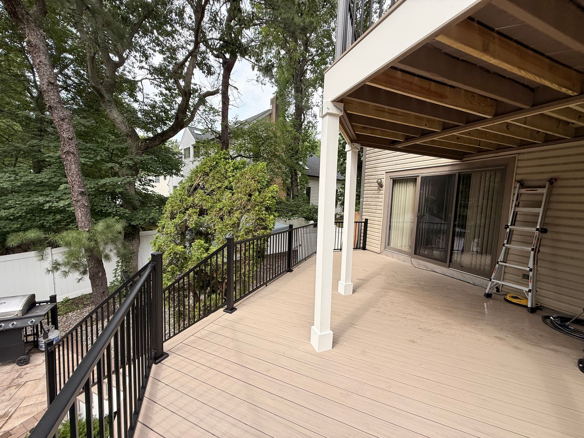 Deck with brown composite decking, black railing, and white support posts.  A ladder leans against the house.
