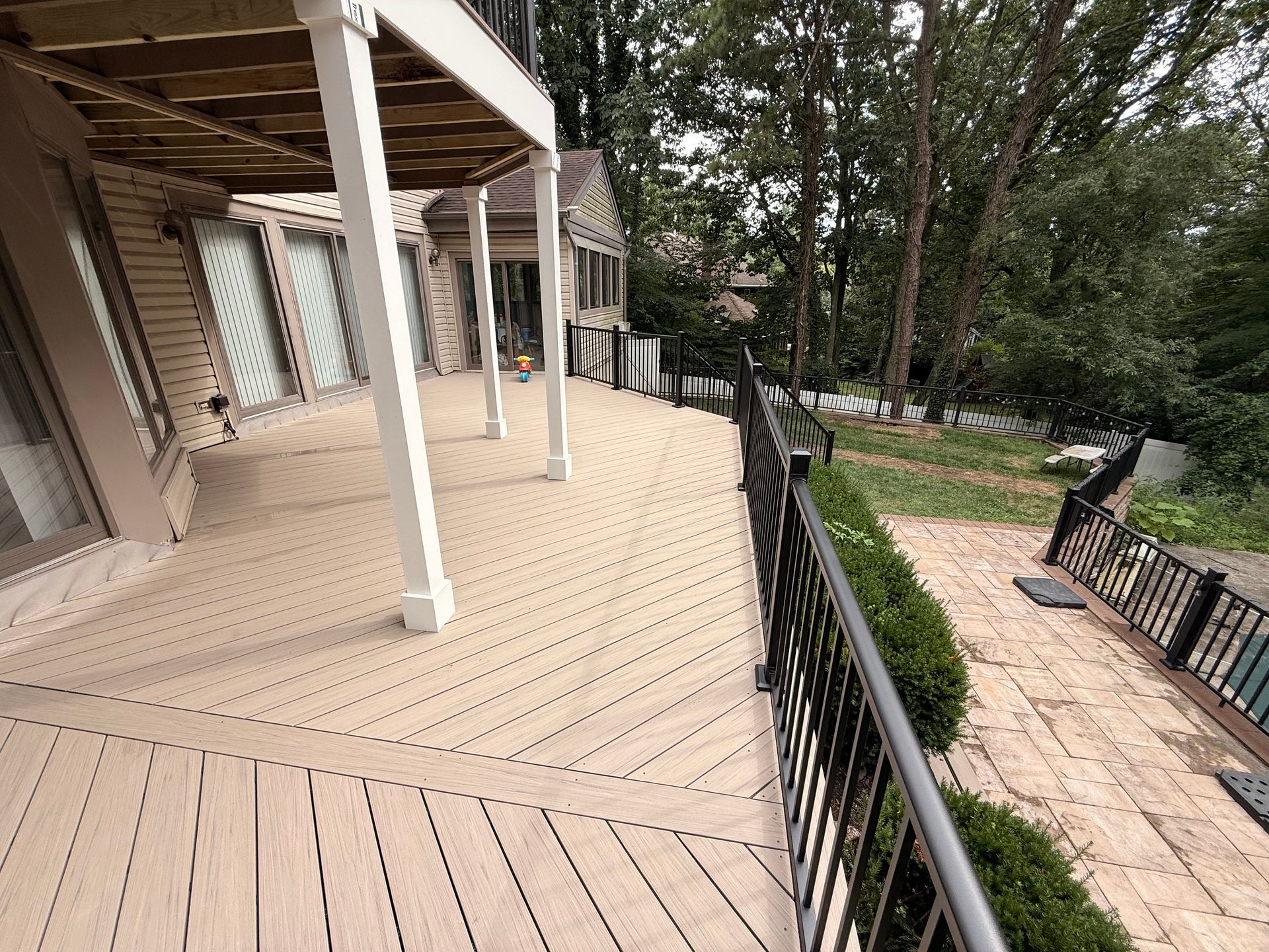 Wooden deck with black railing, overlooking a brick patio and greenery.