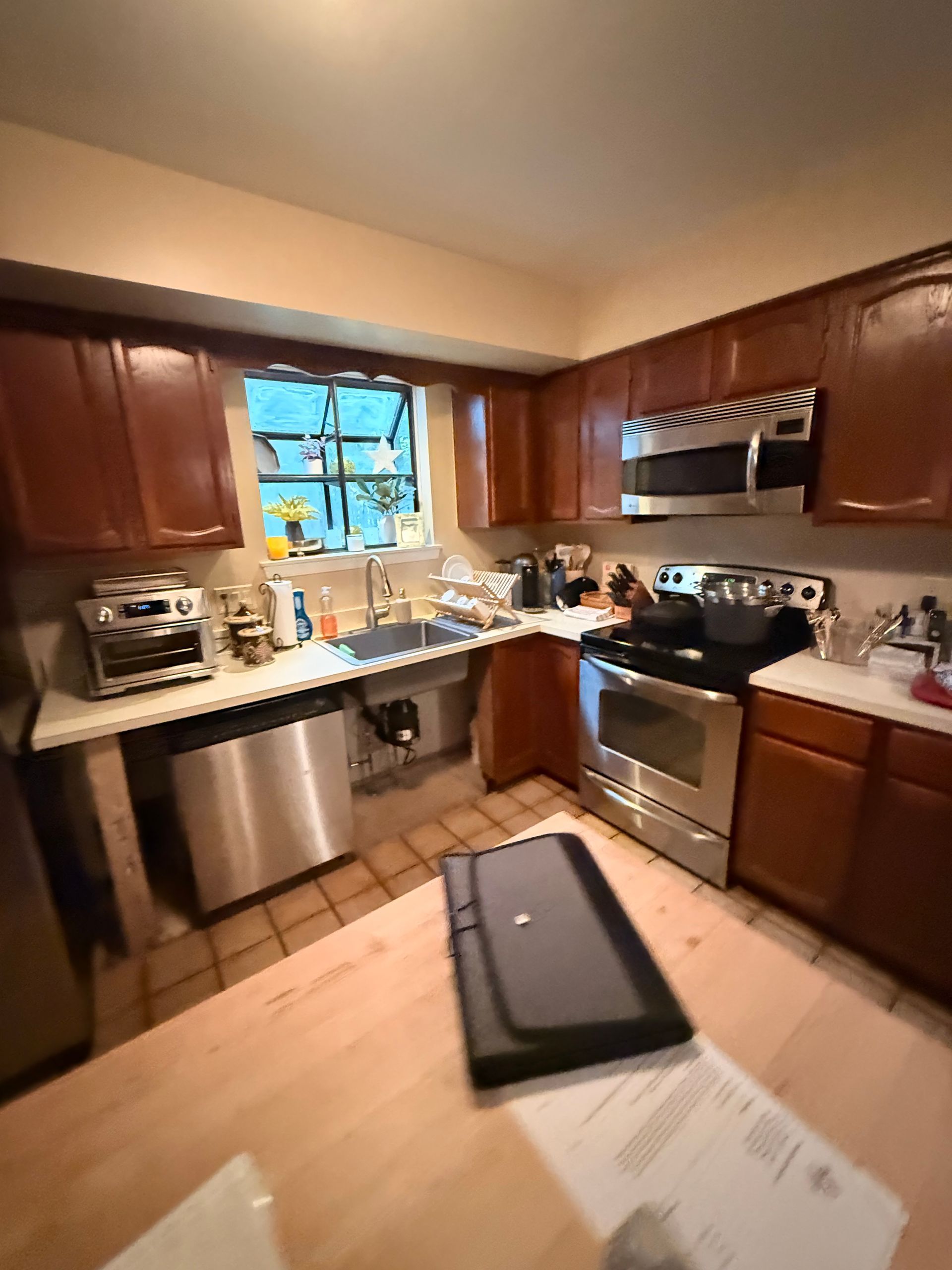 Kitchen with brown cabinets, stainless steel appliances, and a window. A table sits in the foreground.
