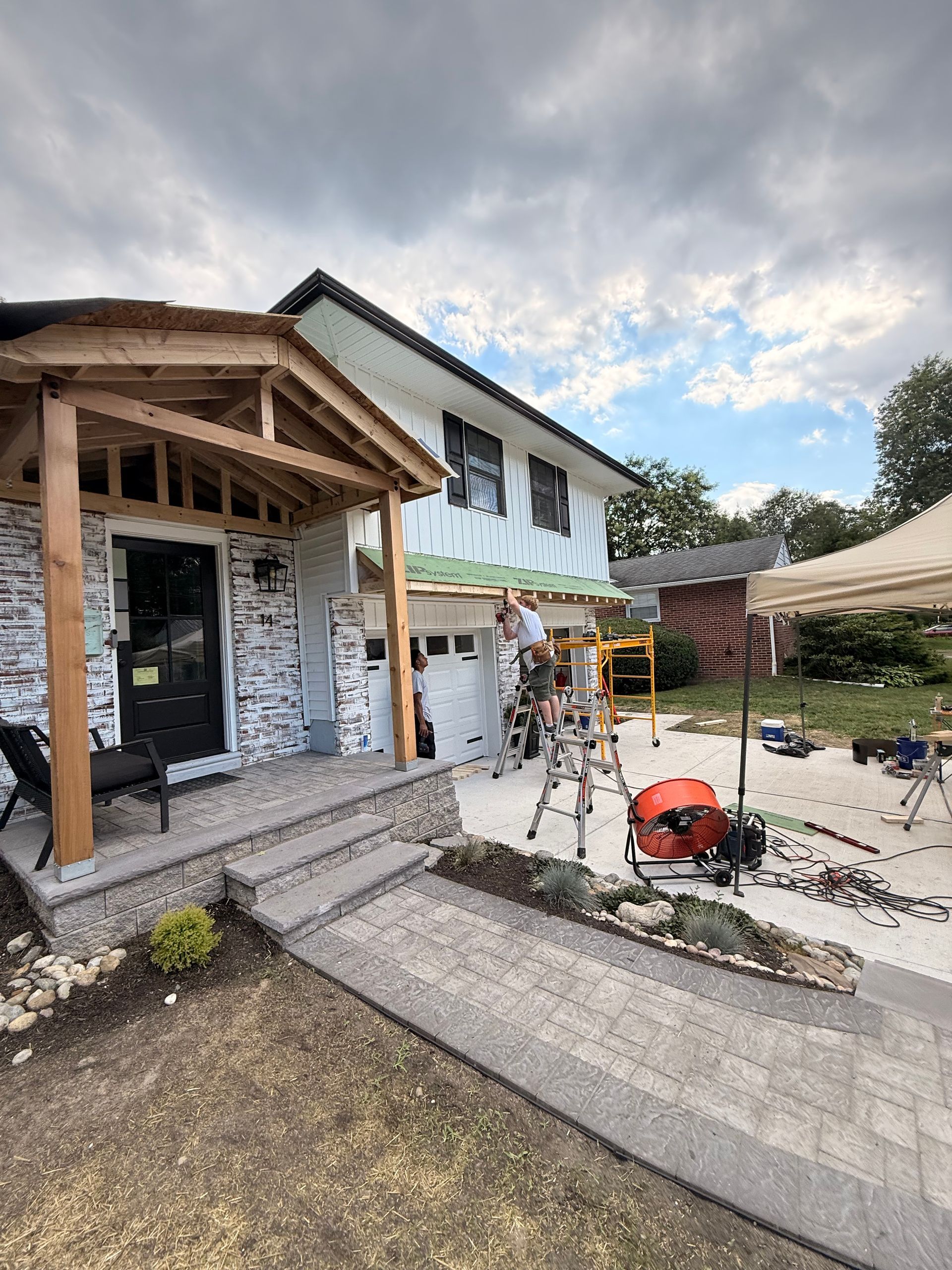 House exterior renovation with workers, ladder, and tools. Grey brick, wooden porch, and cloudy sky.