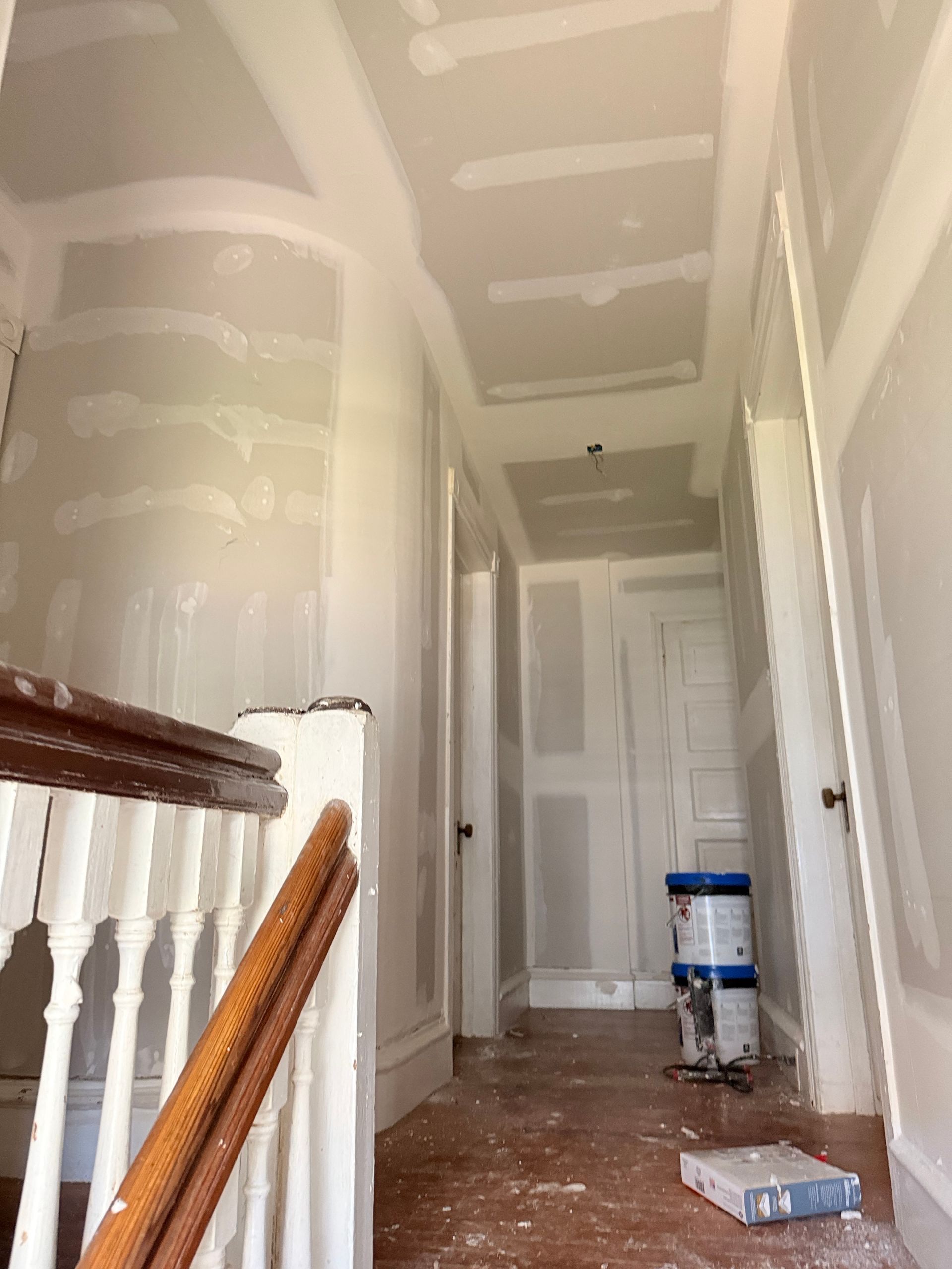 Hallway under construction with drywall and paint buckets visible. A staircase with wooden railing is on the left.