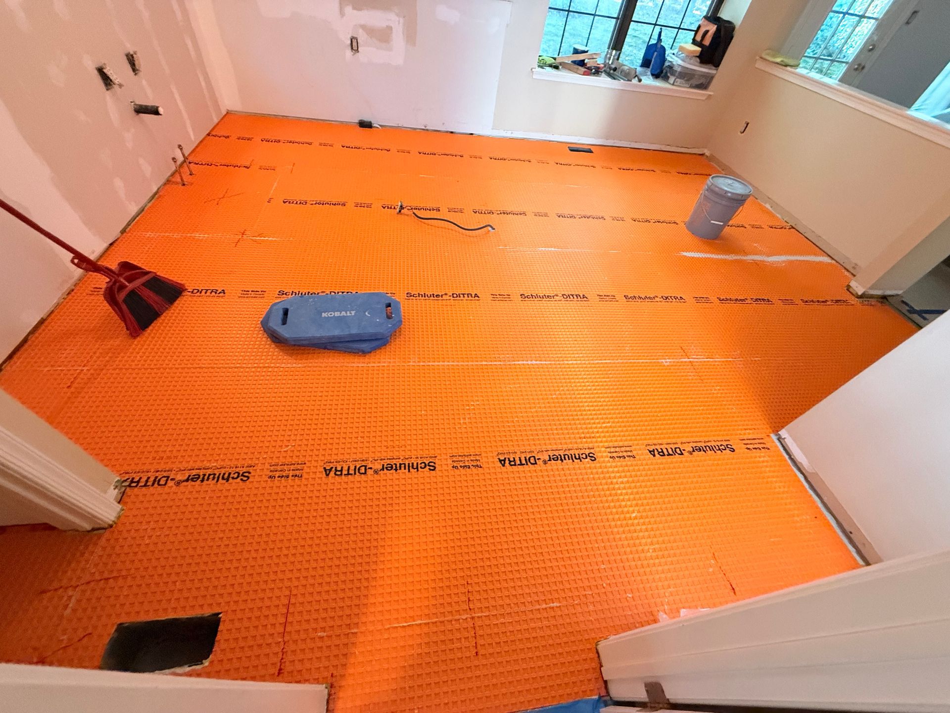 Orange underlayment on a room's floor, ready for tile installation; a broom and tool case are present.