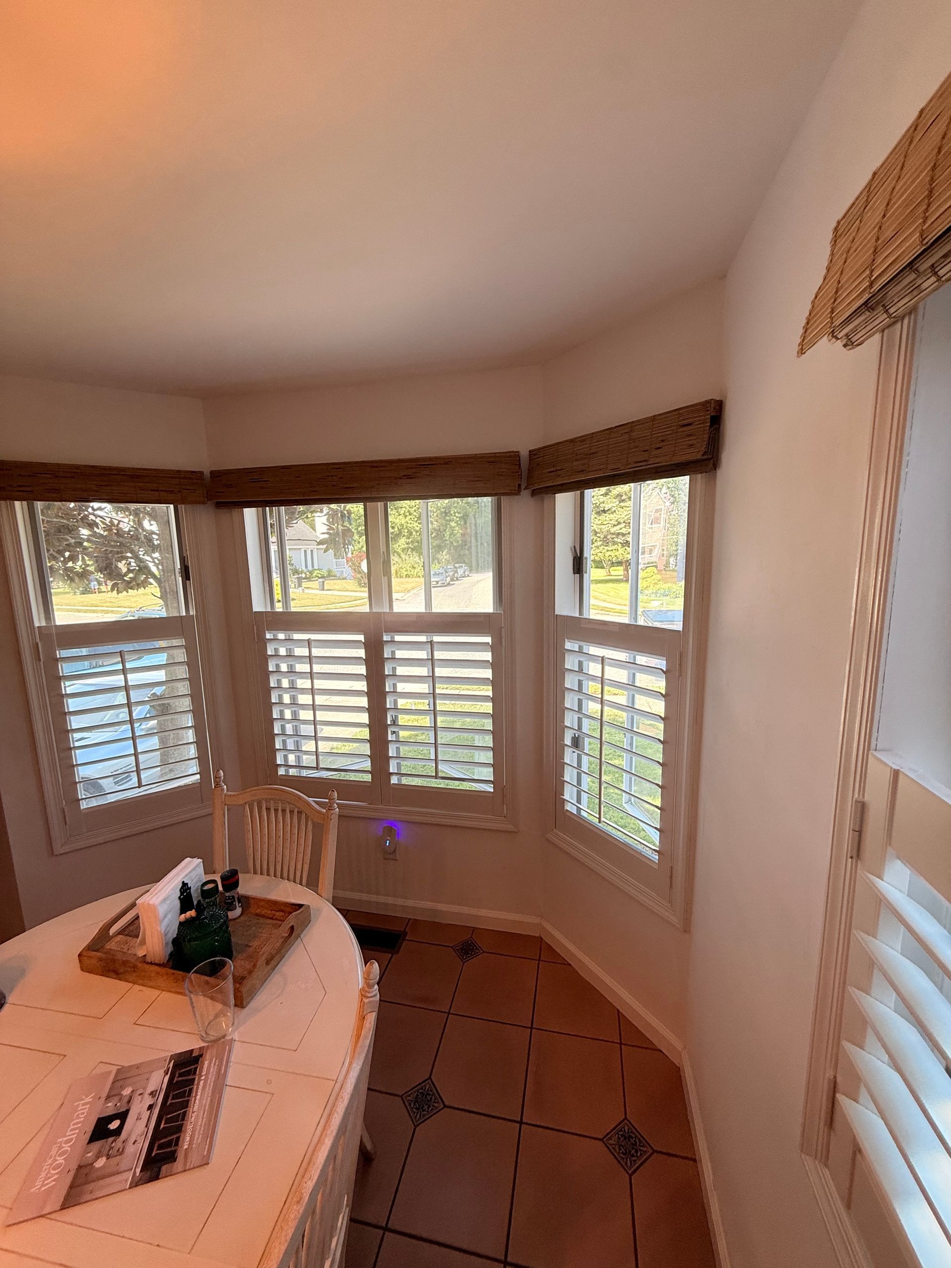 Corner room with windows, shutters, and woven window treatments. Table and chair visible.