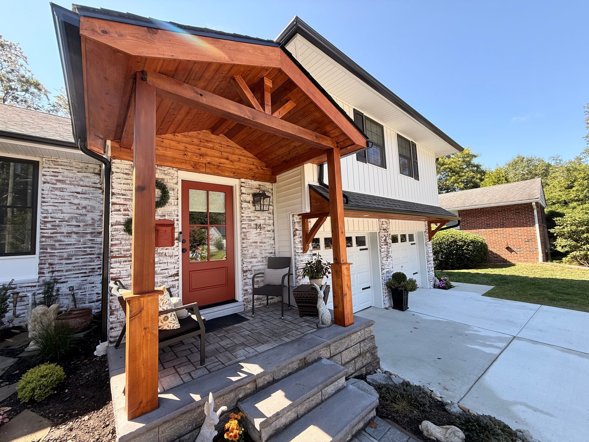Brick home with wooden porch and red door. White siding, two-car garage, and driveway.