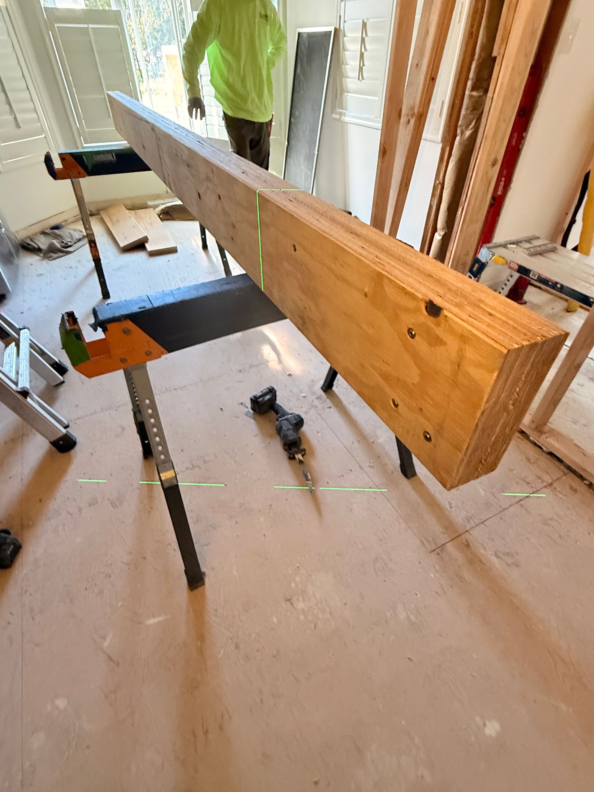 A long wooden beam on a sawhorse in a room under construction. A person stands nearby.