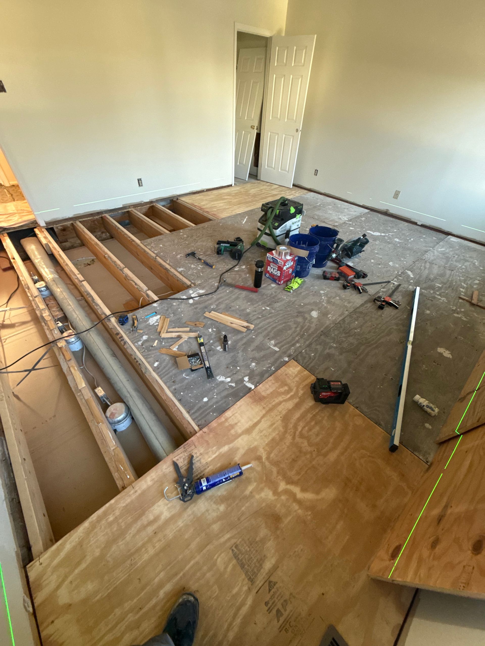 Room under renovation: exposed floor joists, plywood, tools scattered on subfloor, doorway visible.