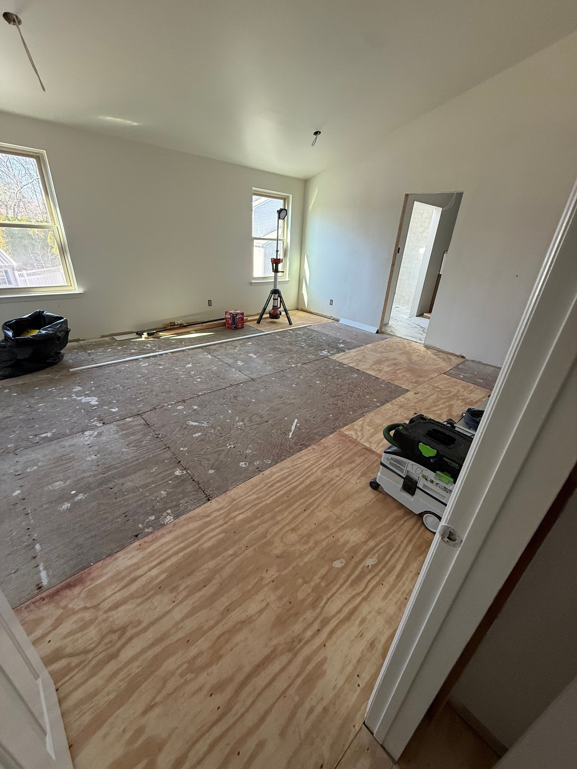 Room under renovation with plywood floor and exposed subfloor. Walls are painted white. Window on the left.