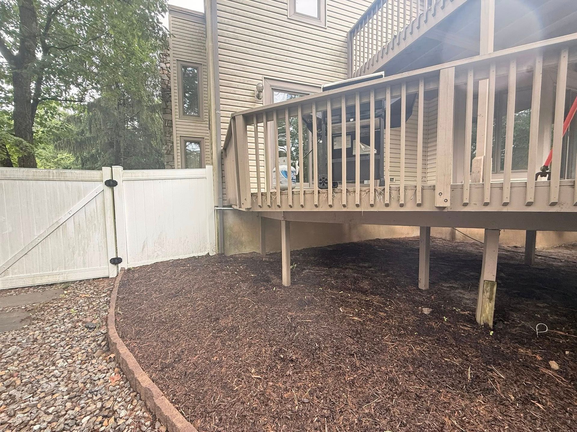 A backyard with a wooden deck, mulch ground cover, and a white fence.