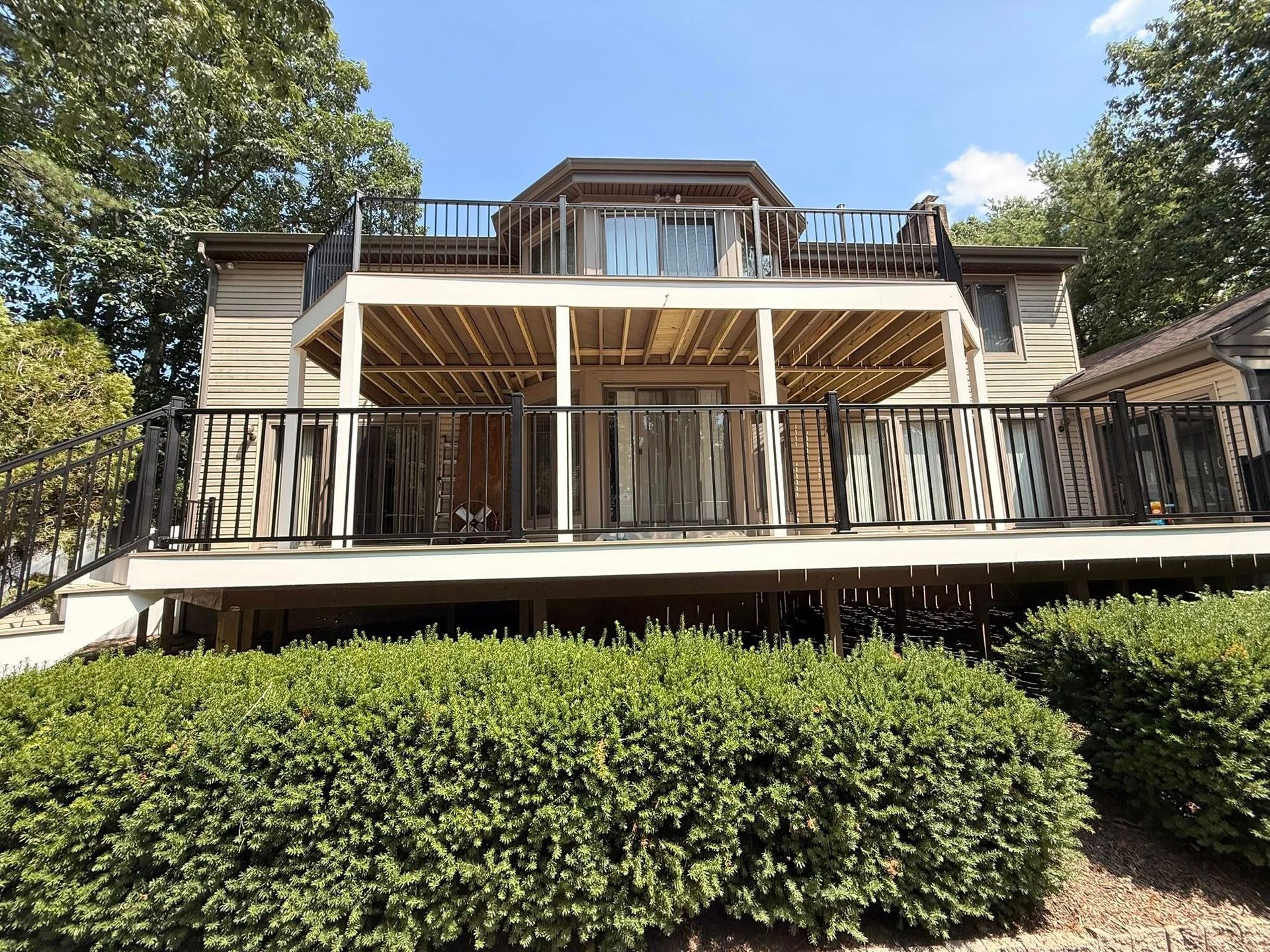 Two-story house with decks, tan siding, and black railings. Green bushes in the foreground.