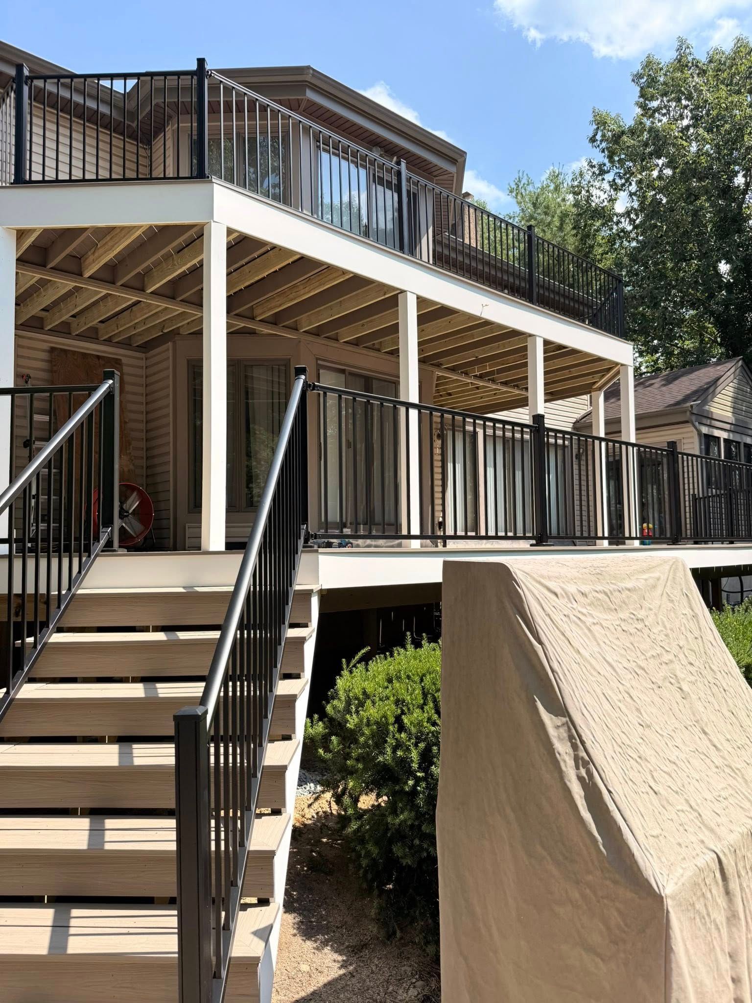 Multi-level deck with black railings and stairs. Overlooks a yard with bushes and a partly visible building.