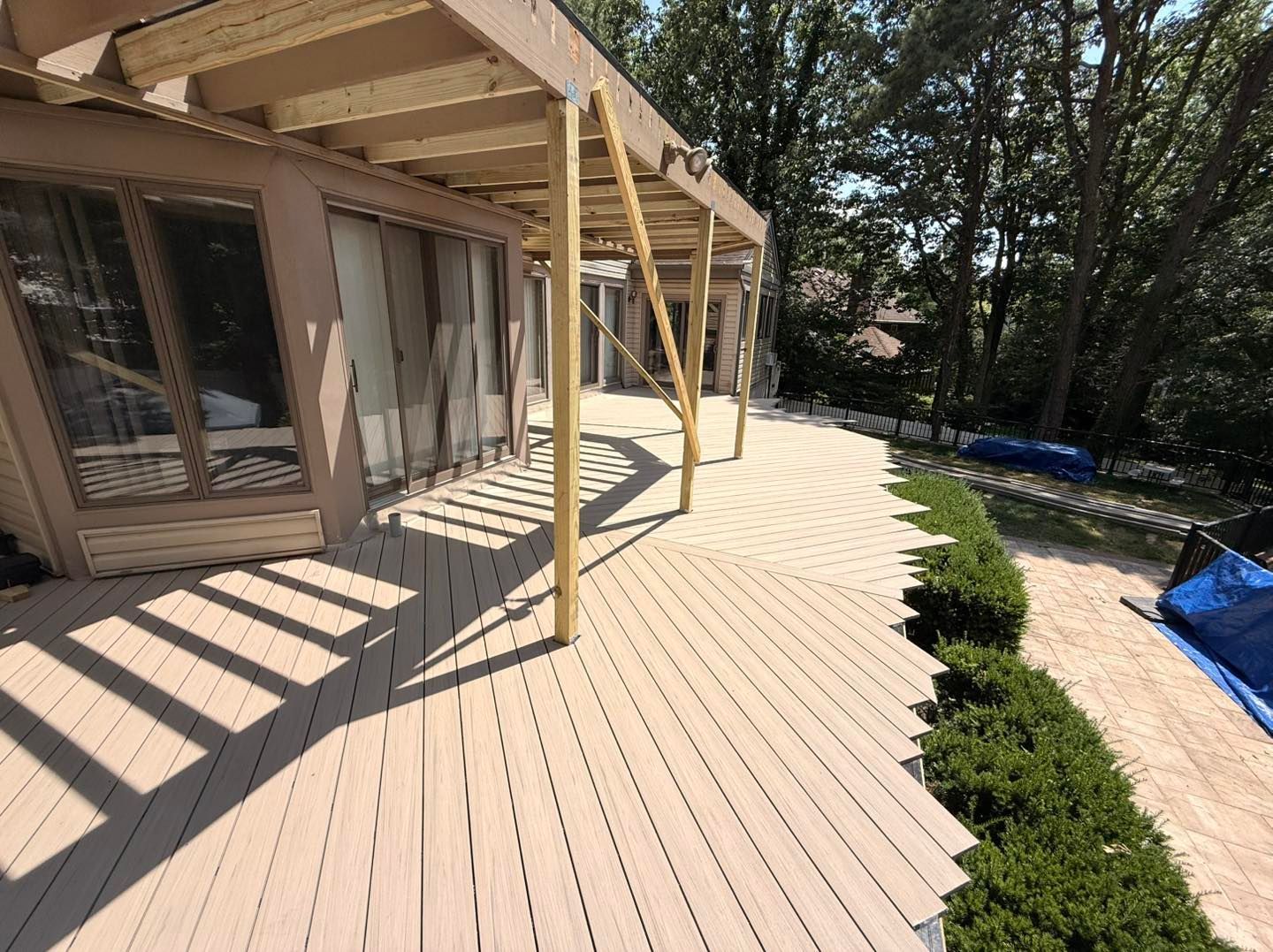 Wooden deck with pergola, beige color. View includes windows, bushes, and trees. Sunny outdoors.