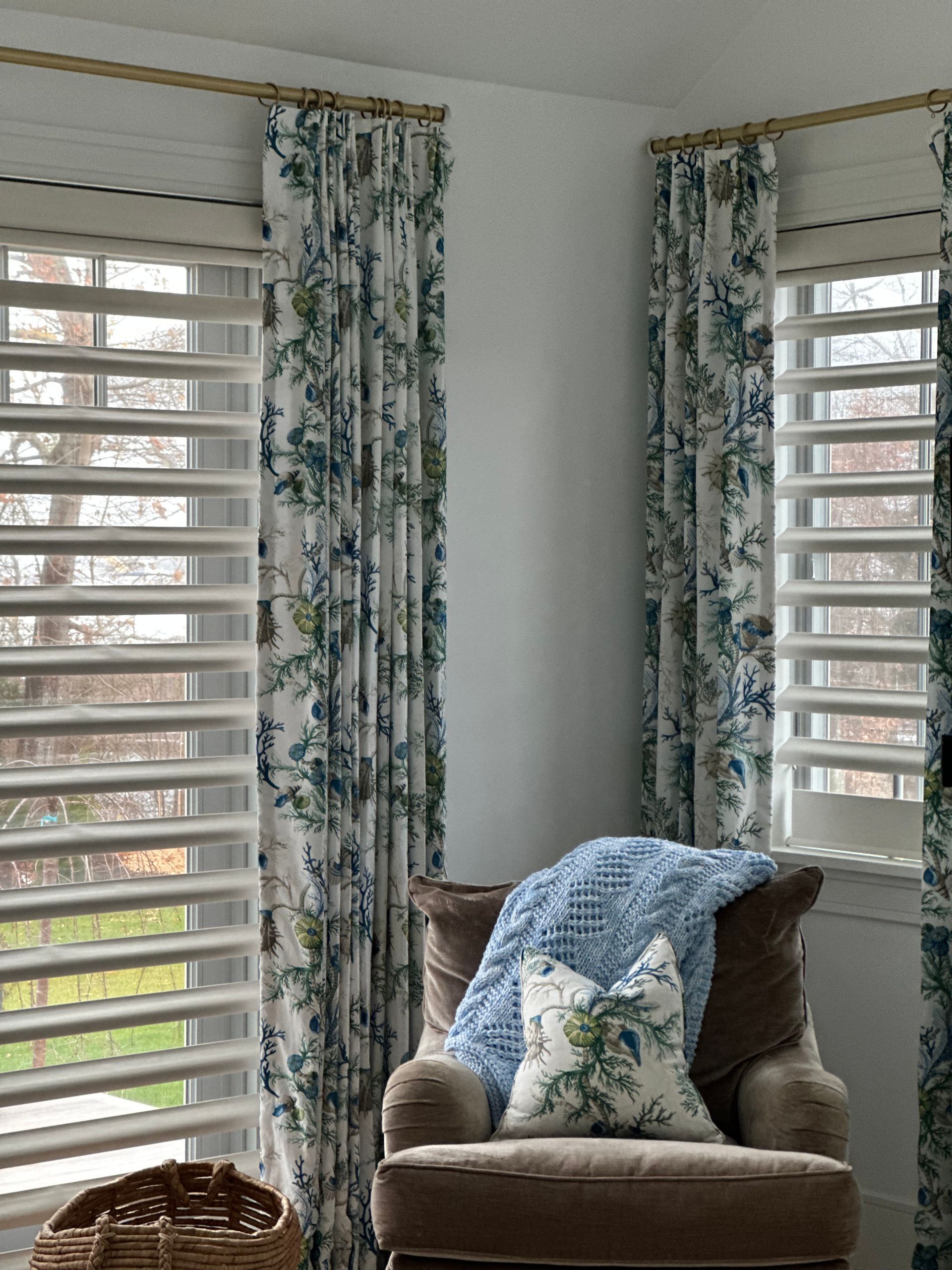 Cozy armchair with blue pillows in a bright room beside two windows with floral curtains and white shutters
