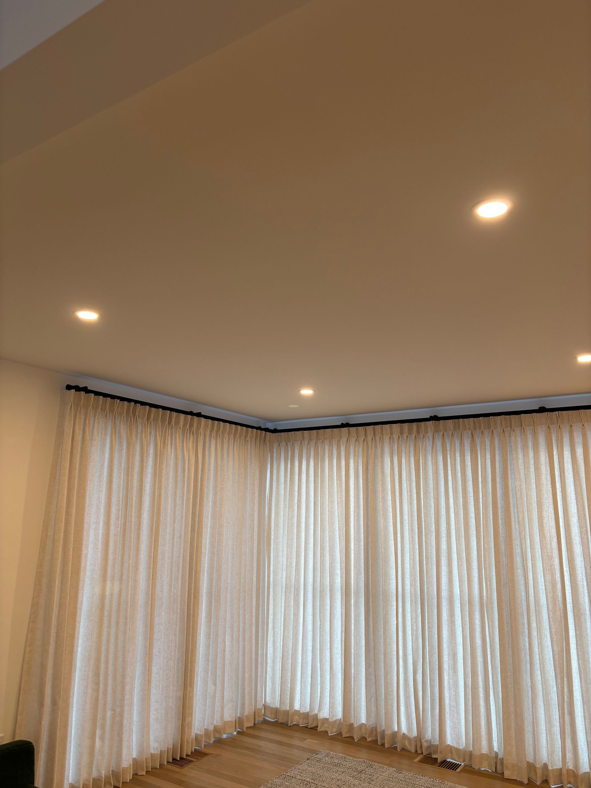 Empty room with beige curtains along two walls, recessed ceiling lights, and a hardwood floor