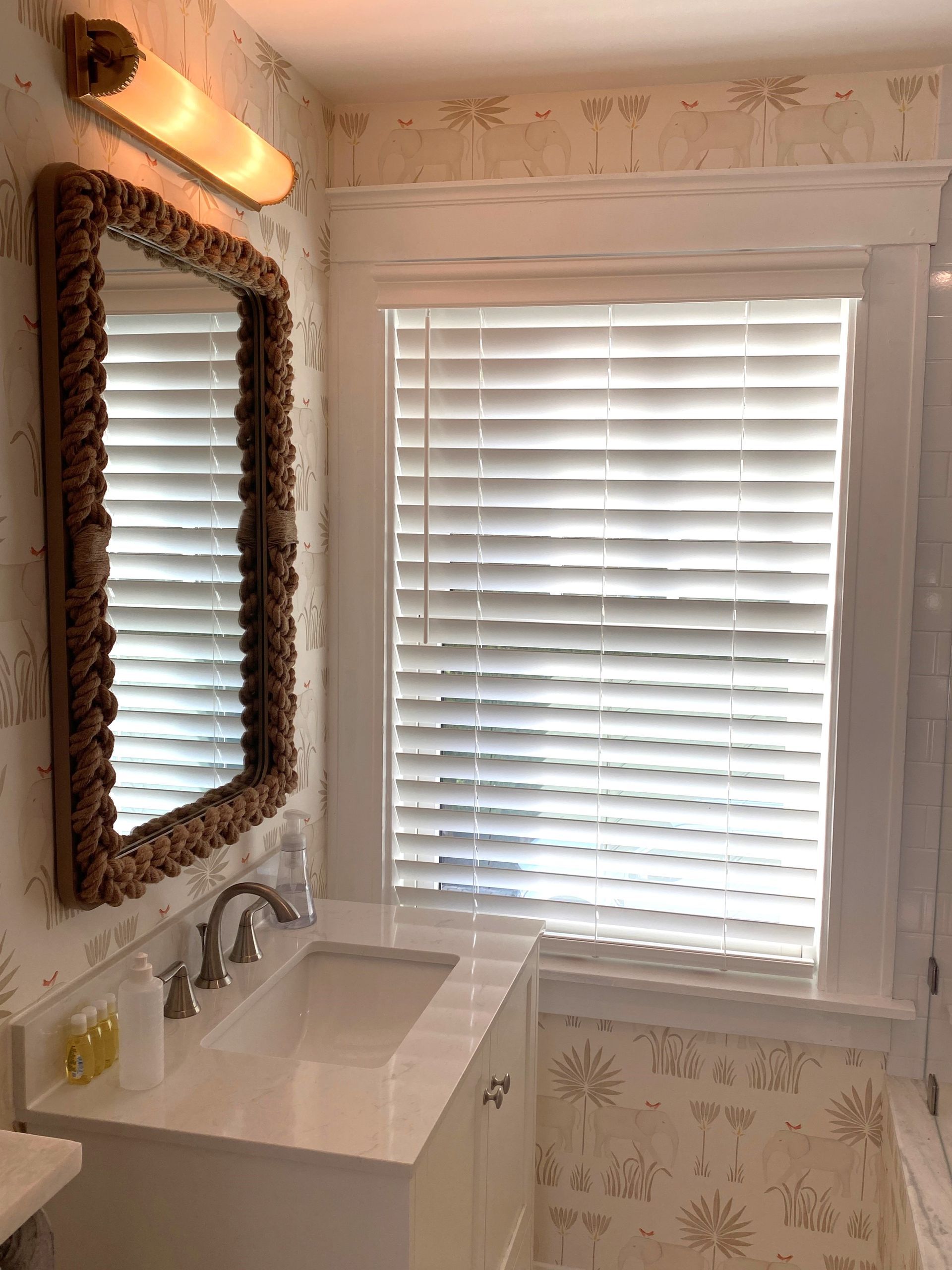 Small white bathroom with sink, mirror, wall light, and window with white blinds.