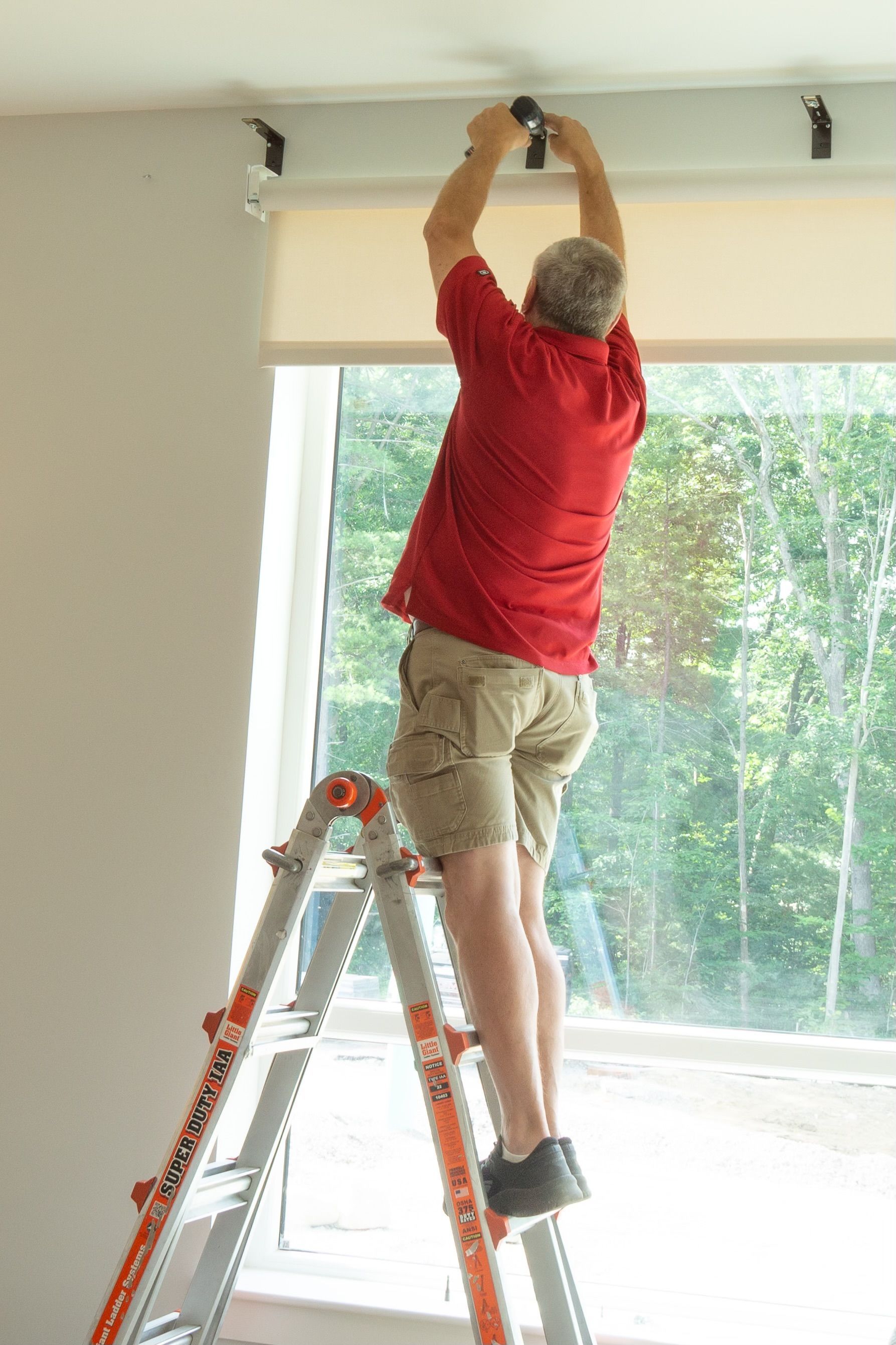 Person on a ladder installing a window blind above a large window.
