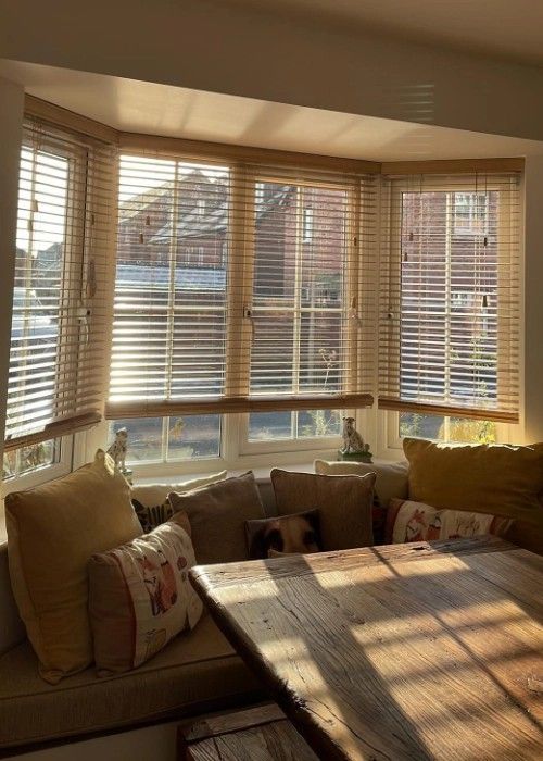 Sunlit living room with bay windows, blinds, and a sectional sofa with pillows and a coffee table