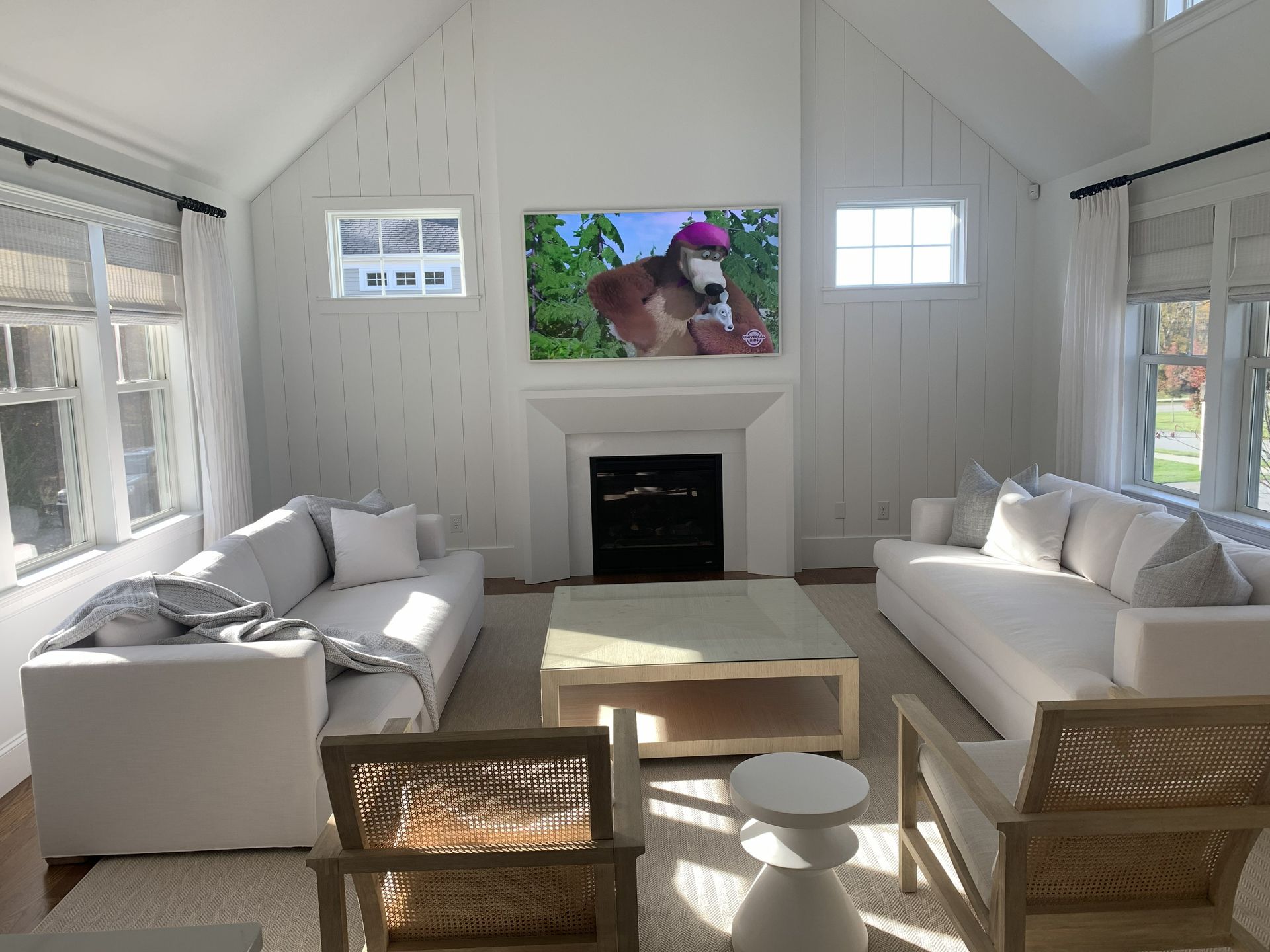 Bright white living room with two sofas, a fireplace, and a TV above it, with sunlight streaming through windows