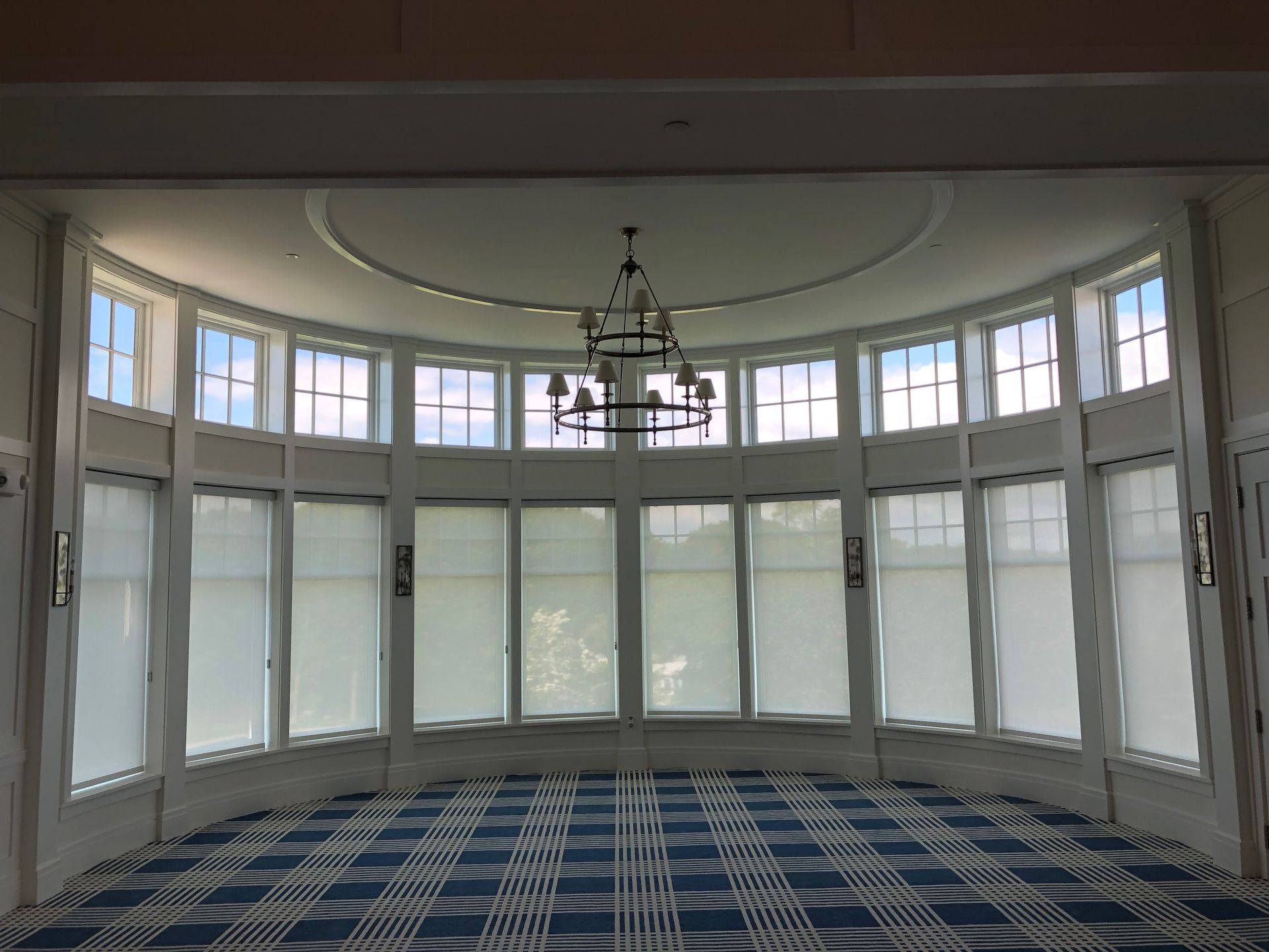 Empty curved room with tall windows, white blinds, chandelier, and blue checkered carpet.