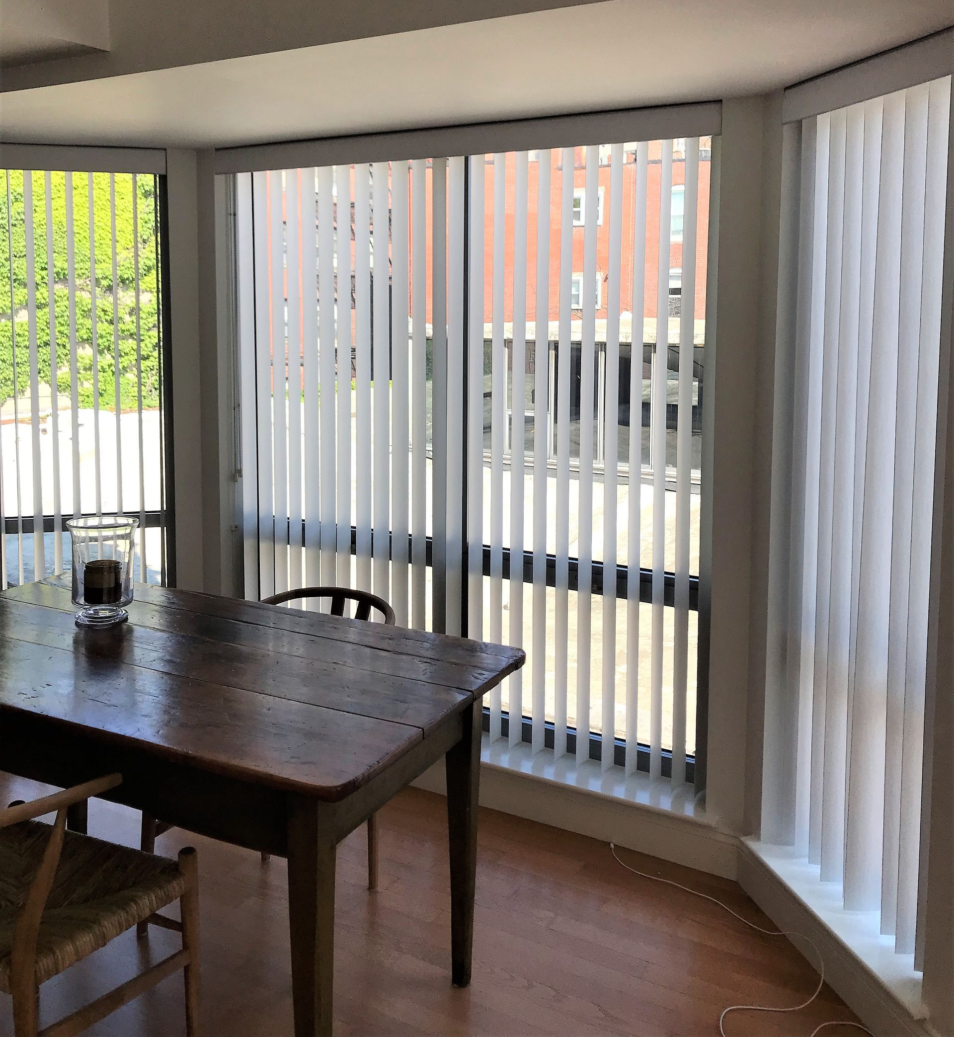 Dining area with dark wooden table beside large vertical blinds overlooking a street outside.