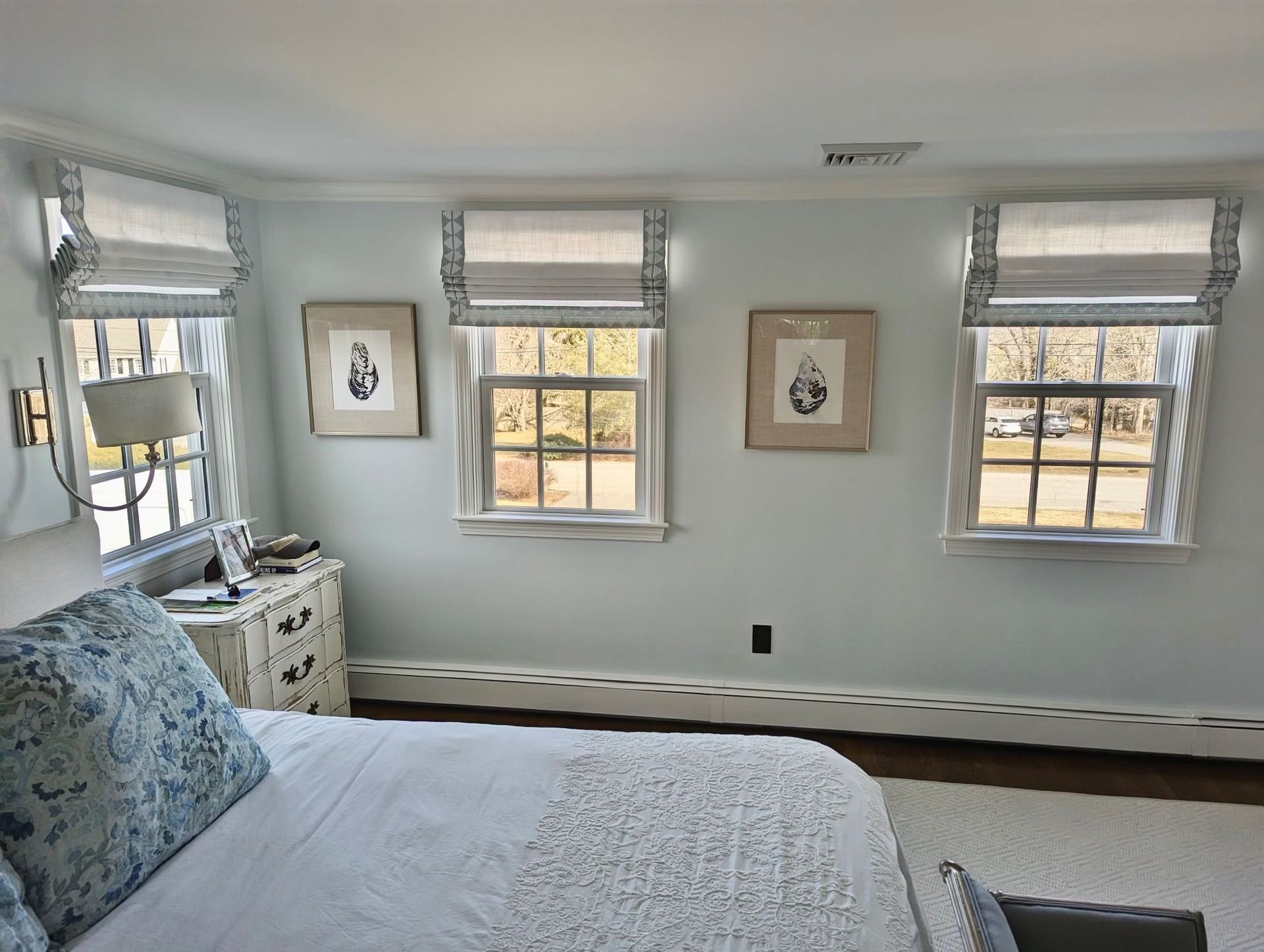 Bright bedroom with white walls, blue bedspread, dresser, and three windows letting in daylight.