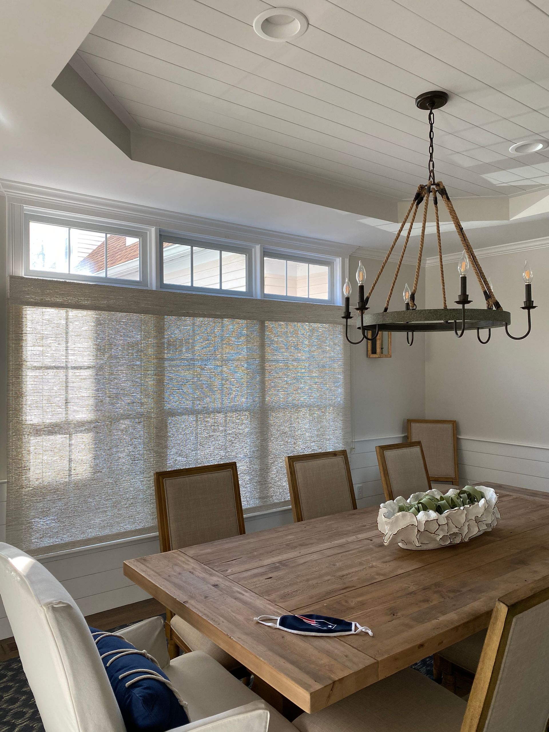 Dining room with wooden table, upholstered chairs, chandelier, and large windows with blinds.