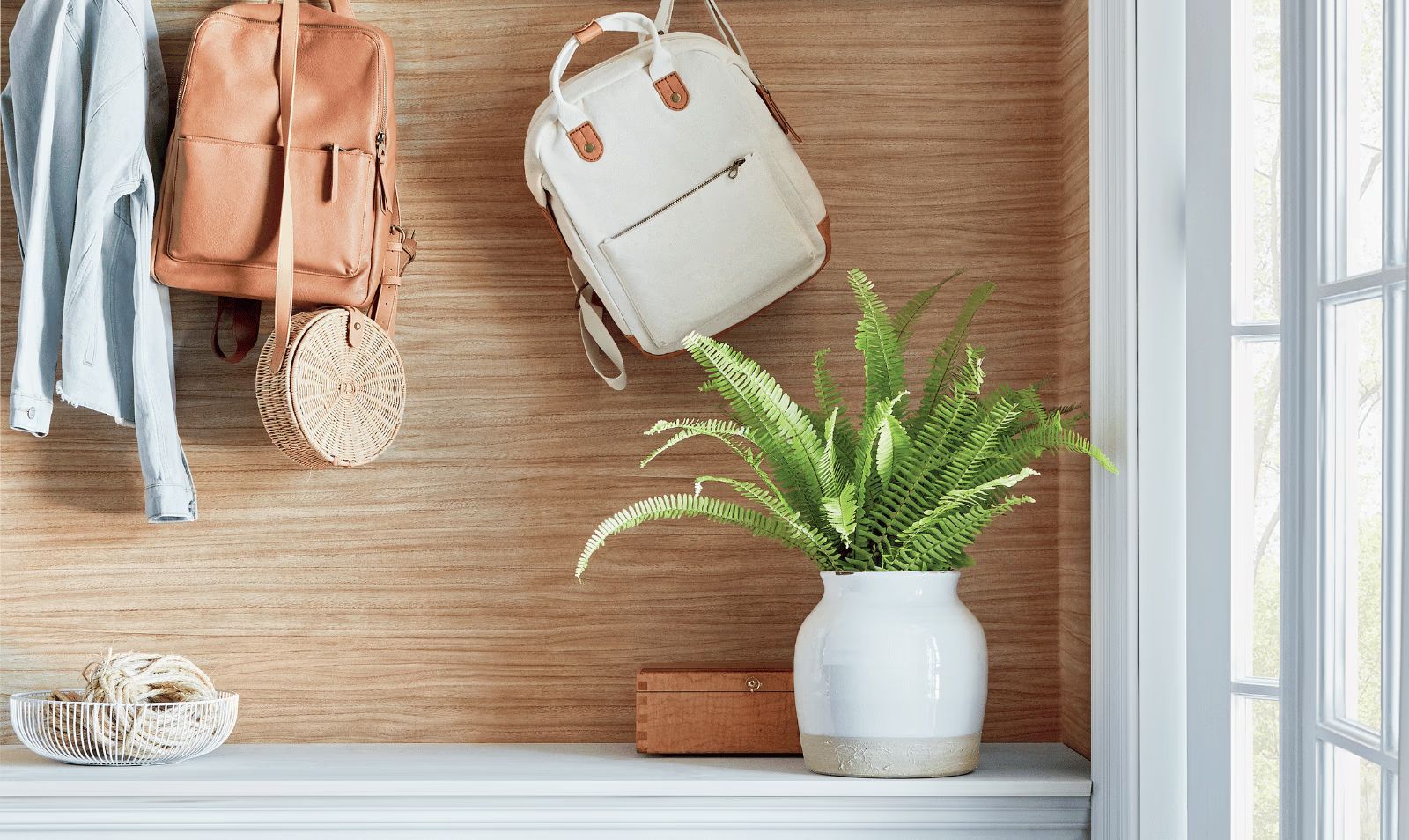 White and tan backpacks hanging on a wooden wall shelf with a potted green plant and a bright window
